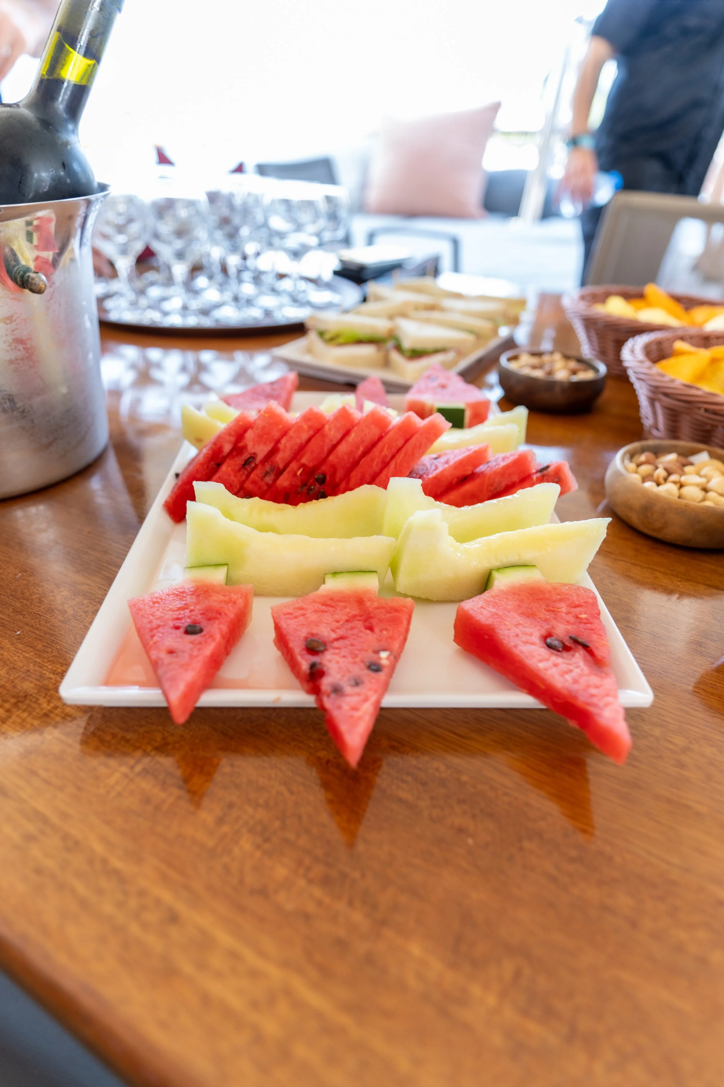 Assorted sliced watermelons on a white rectangular platter on a wooden table, with other food items and glasses in the background.