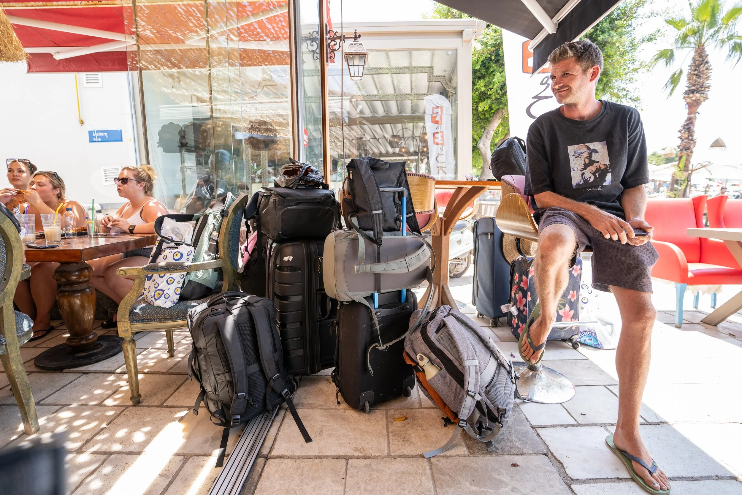 Group of travel backpacks and luggage stacked outside a cafe, with a man sitting on a stool on the right, and four women sitting at a table on the left, enjoying drinks.
