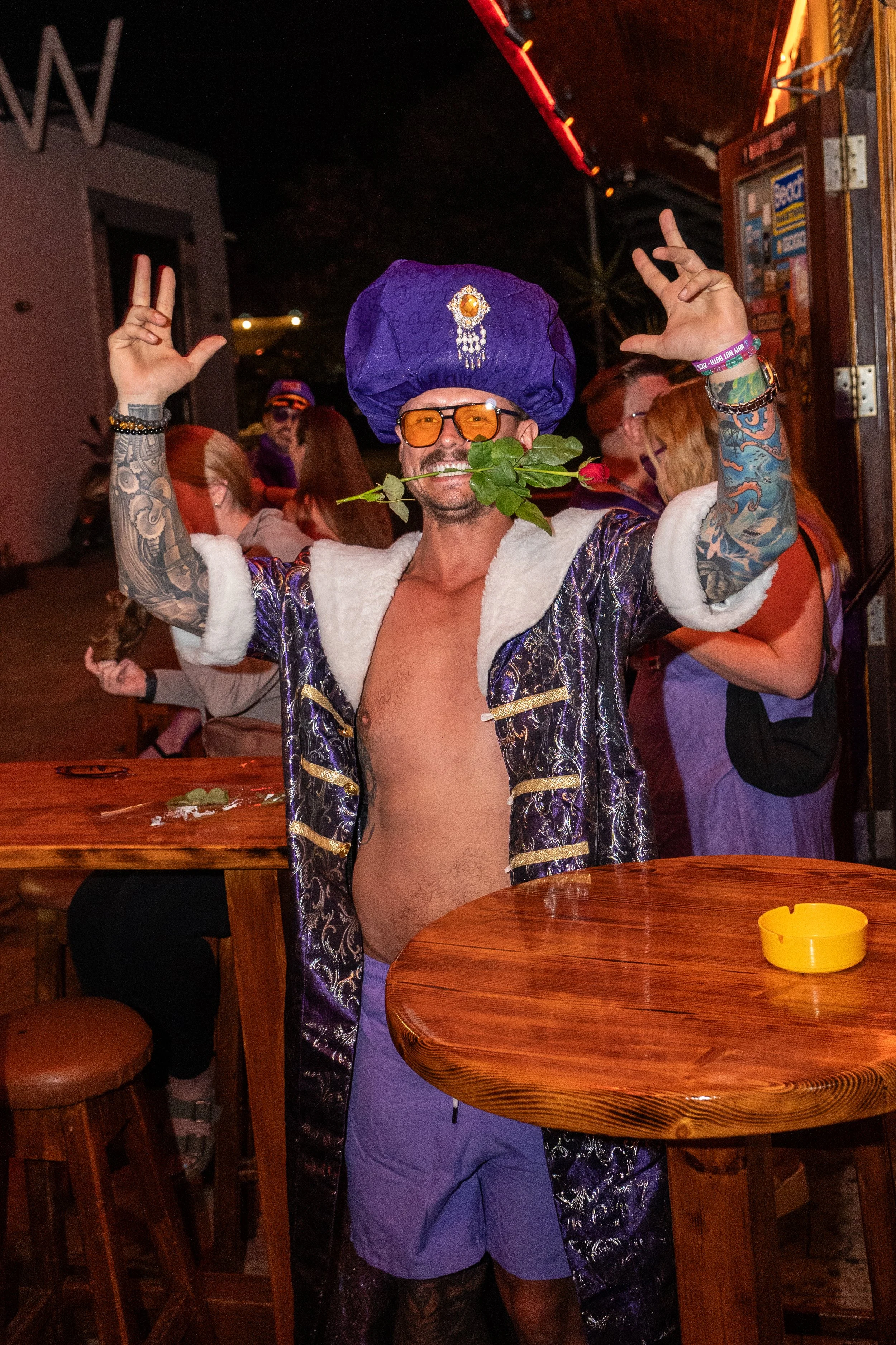Man dressed in a purple sequined coat with white fur trim, purple shorts, wearing an elaborate purple turban with gold and jewel embellishments, orange-tinted glasses, and holding a rose in his mouth, smiling at a lively party.