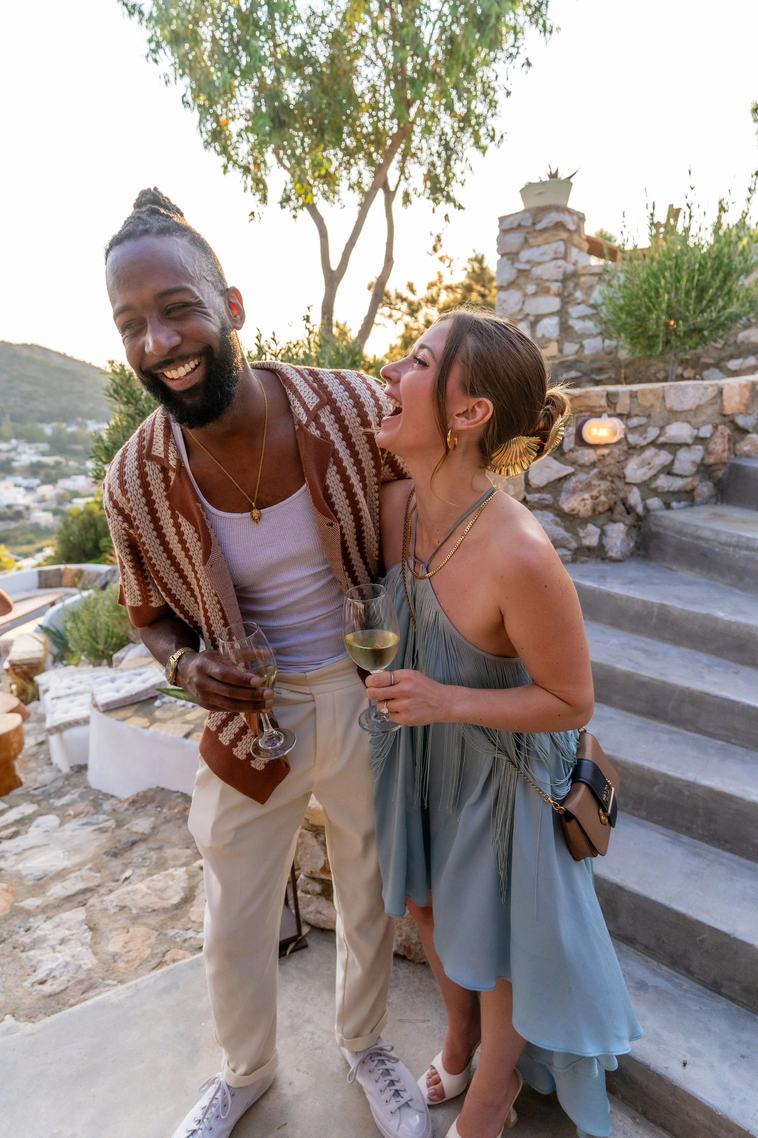 A man and woman are laughing and holding glasses of white wine outdoors during sunset, with stairs and a stone wall in the background.