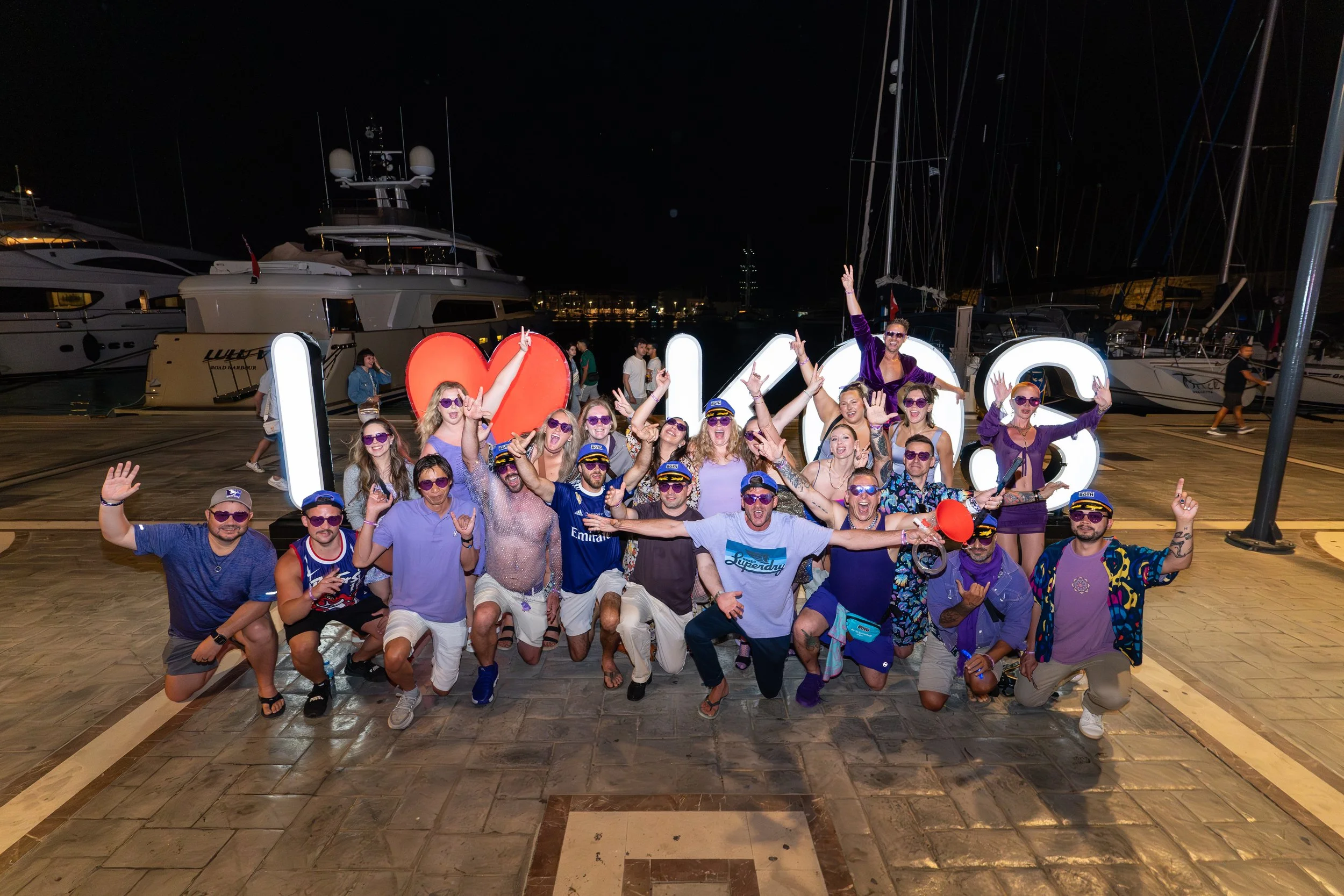 Group of people wearing purple sunglasses at night in front of illuminated letters spelling 'I ❤️'. They are on a dock with boats in the background, celebrating and smiling.