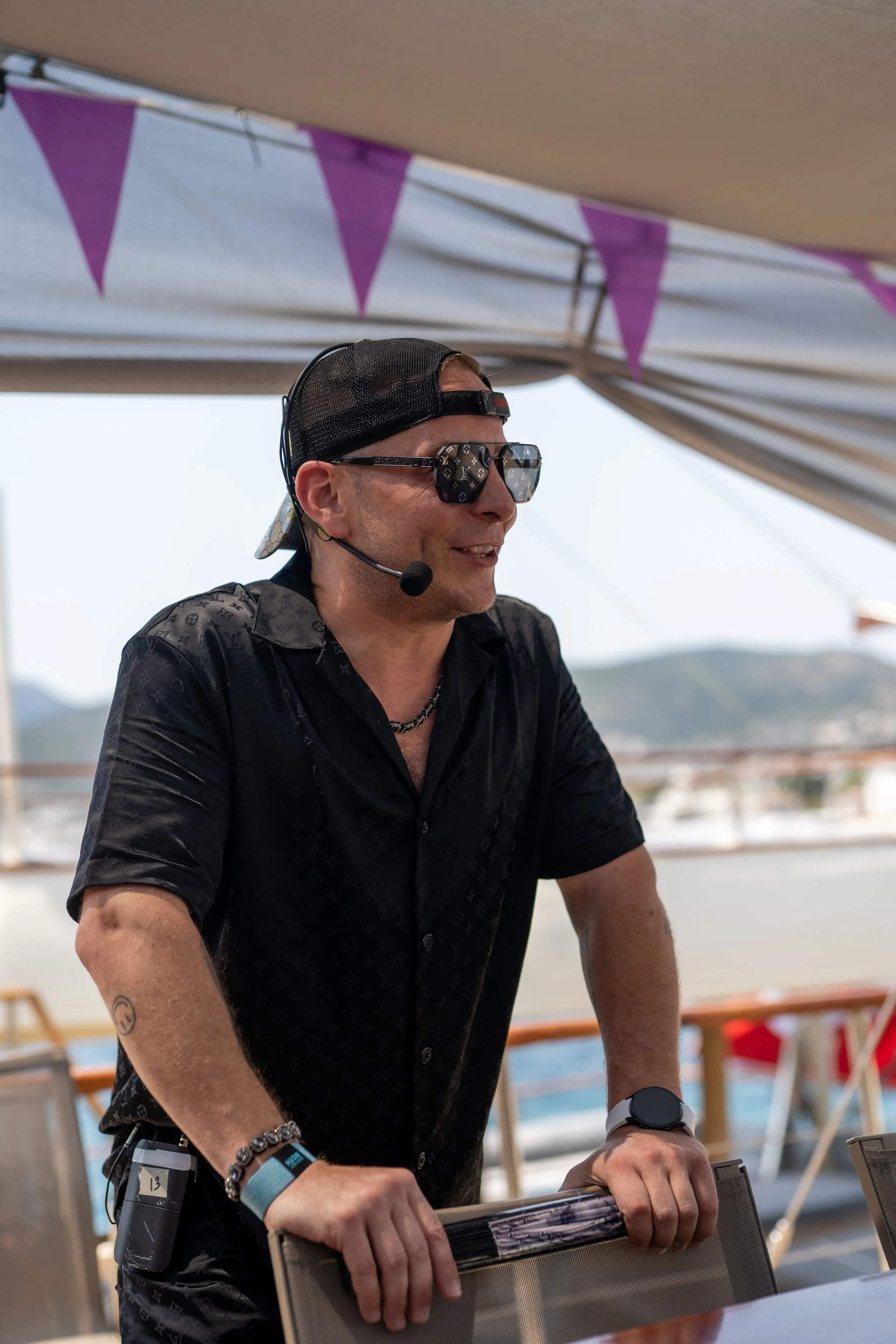 A man wearing sunglasses, a black cap, and a black shirt with a Louis Vuitton pattern, standing outdoors under a canopy with purple bunting, smiling and holding onto a chair.