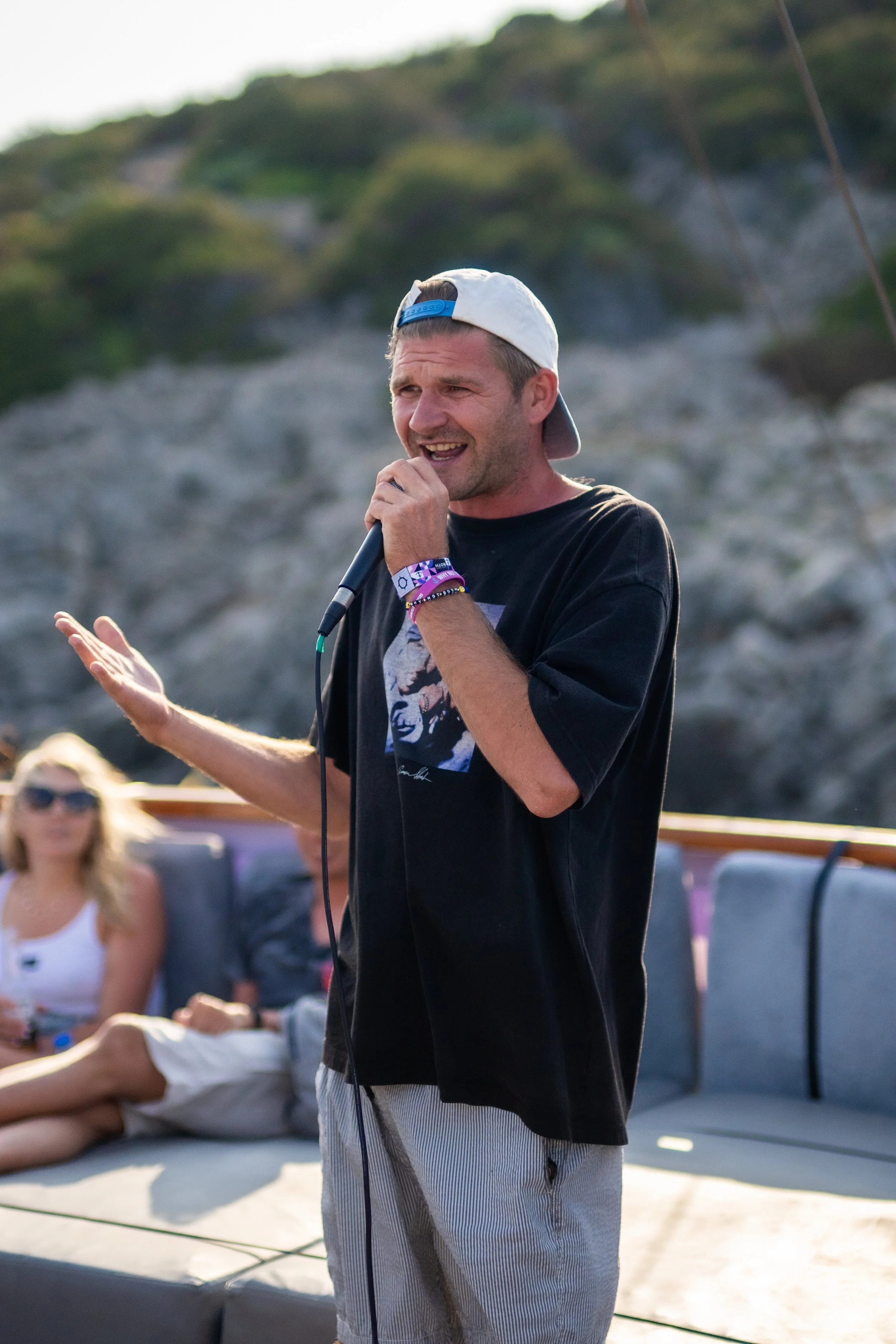 Man with a backward cap singing or speaking into a microphone on a boat with a woman sitting in the background.