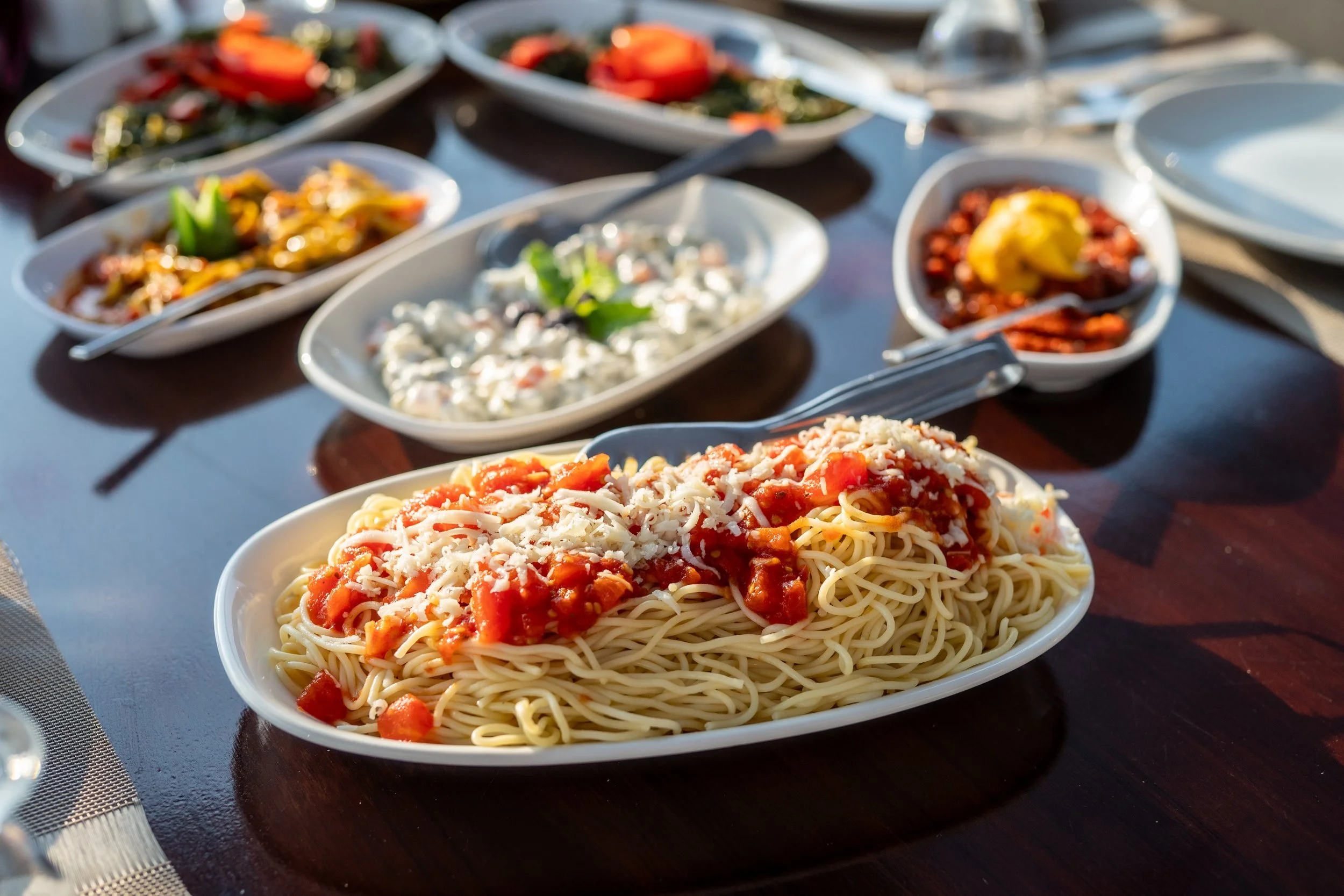 Spaghetti with tomato sauce topped with shredded cheese, surrounded by various salads in oval and round bowls on a wooden table.