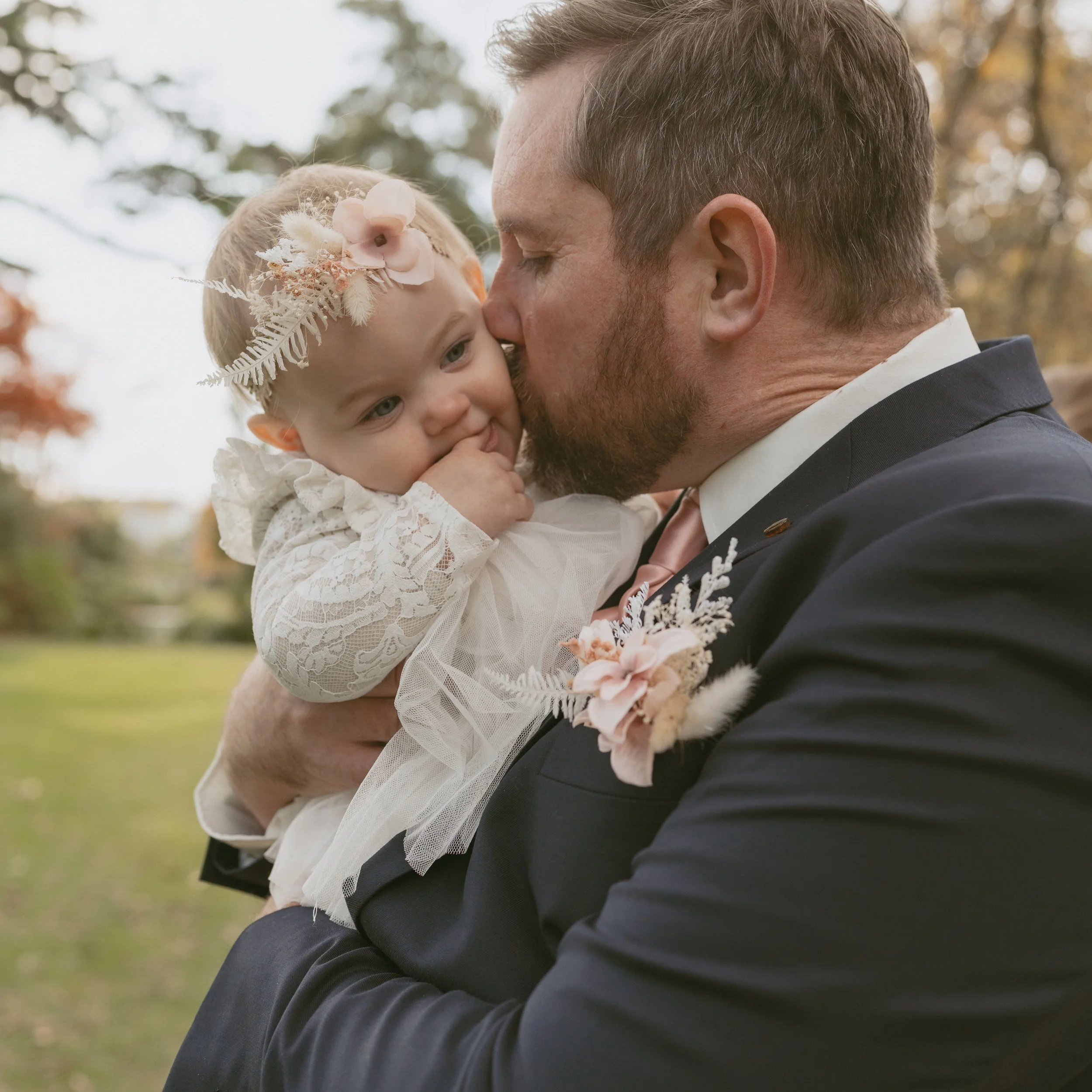 Groom wearing suit and pocket square boutonnière, holding and kissing baby daughter who is wearing a flower crown