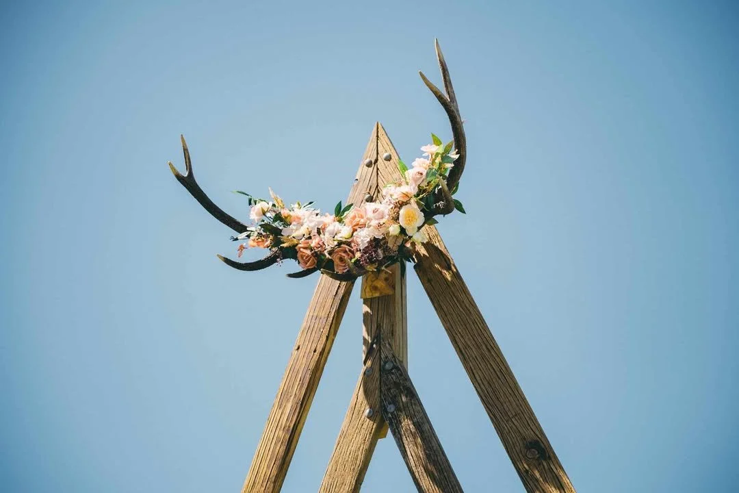 Decorative wooden teepee with antlers adorned with pink and white flowers against a blue sky.