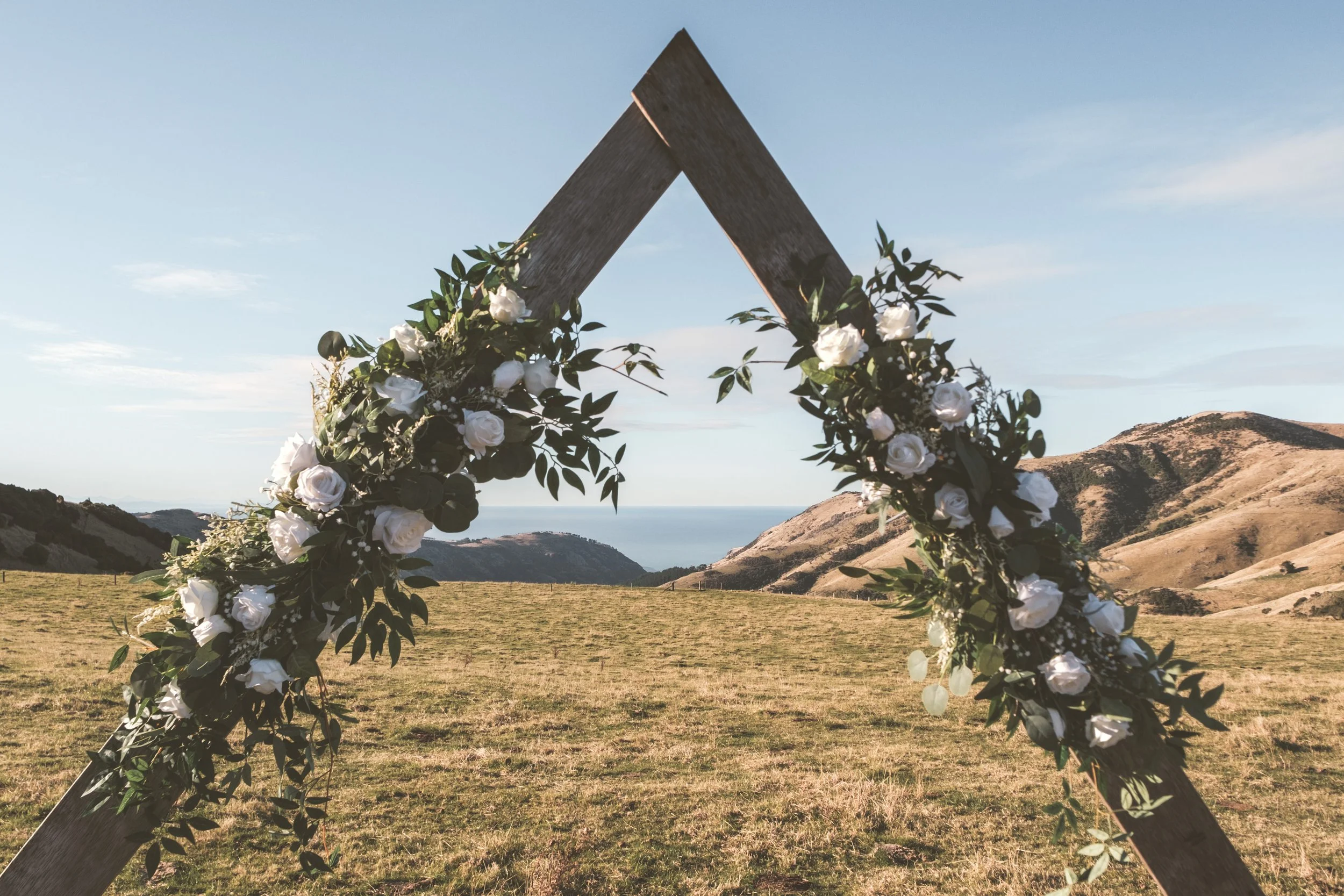 Wooden arch decorated with white roses and greenery on a grassy field with hills and mountains in the background under a blue sky.