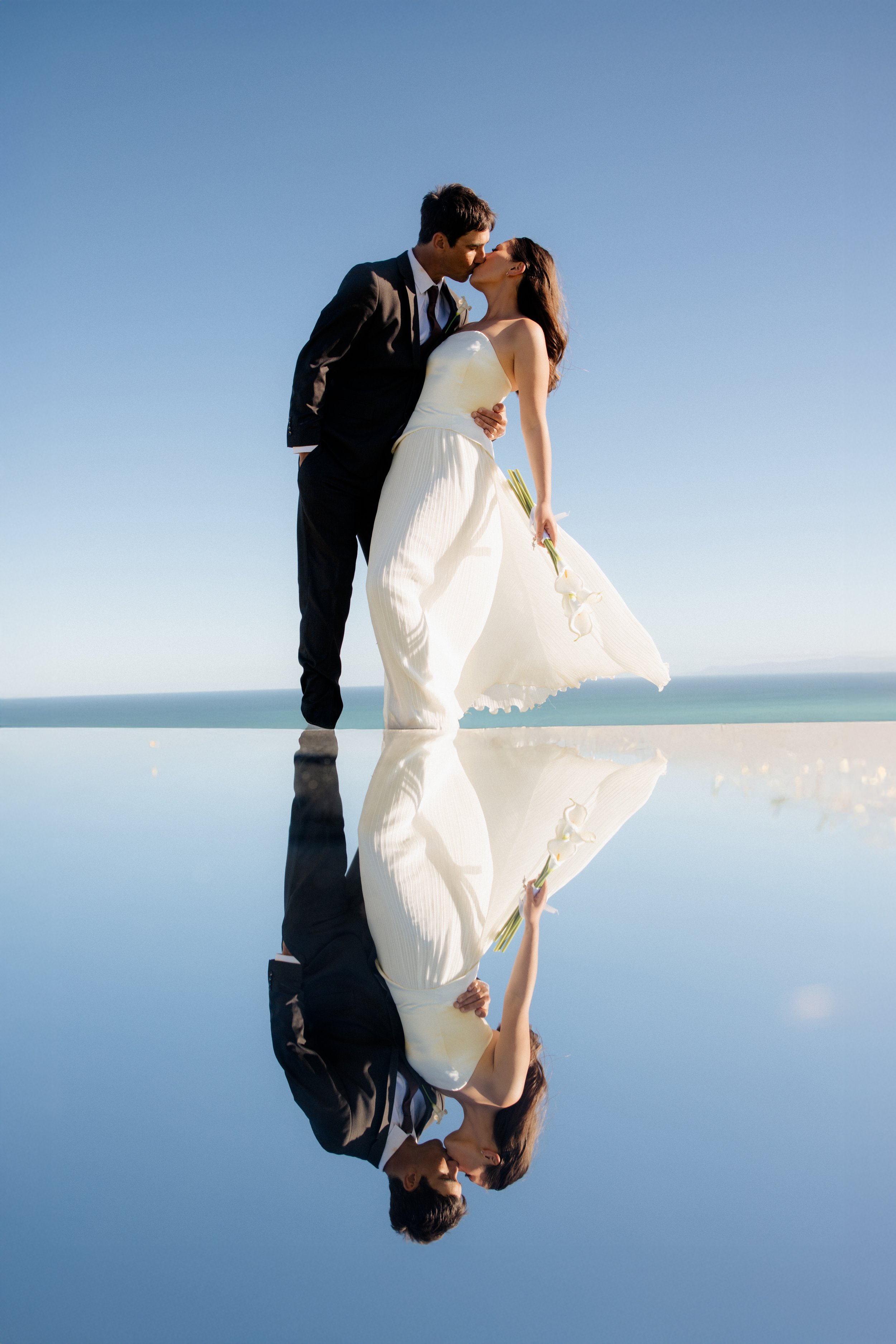 stunning photo of bride and groom kissing with a mirror in the foreground for that reflection effect, bright blue skies and bride holding calla lily bouquet