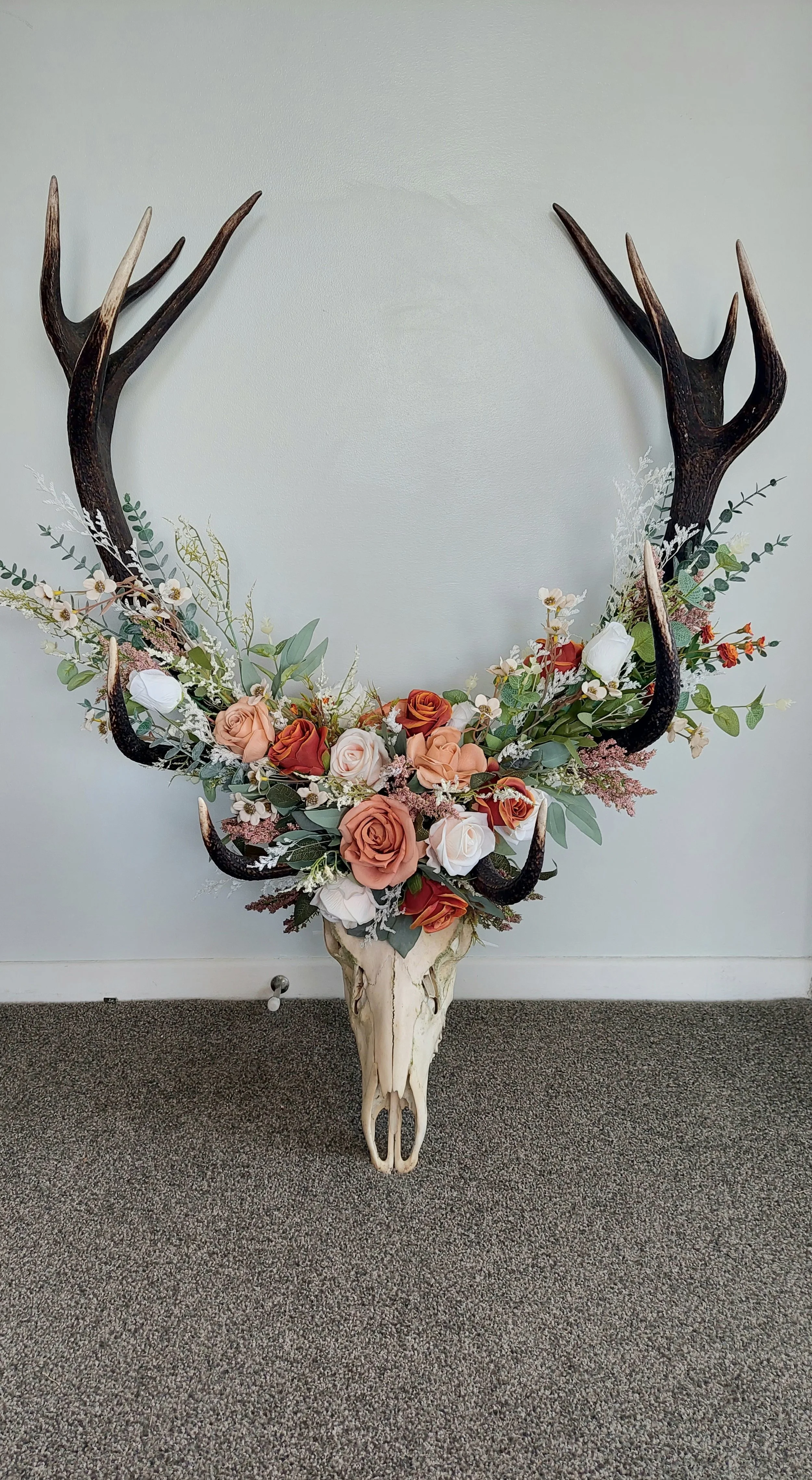 Decorative wall piece featuring a deer skull with antlers, adorned with flowers and greenery.