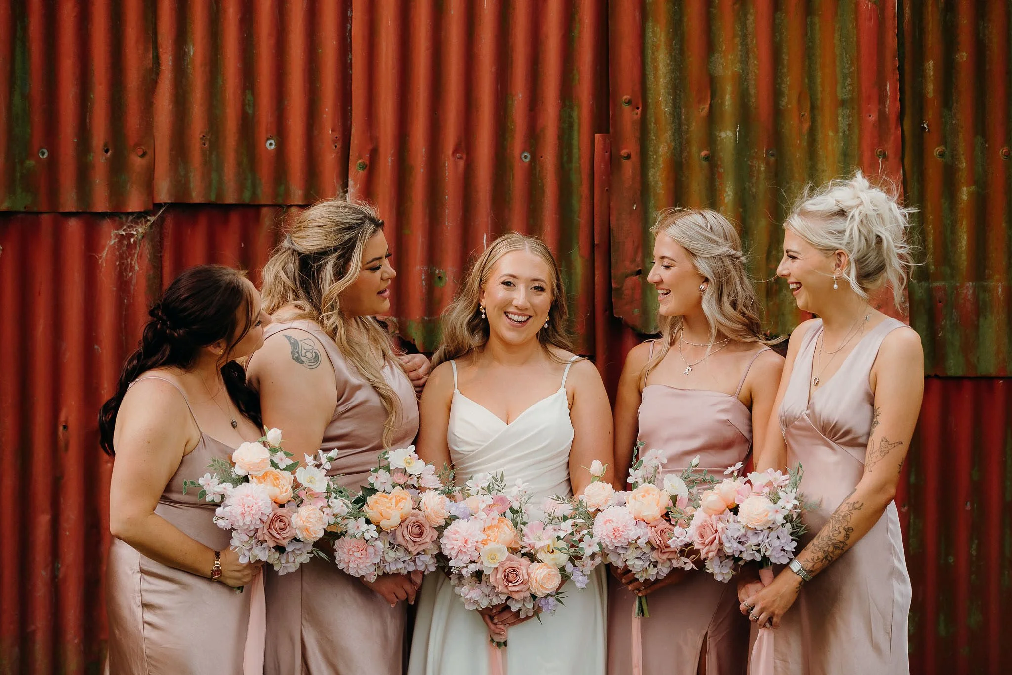 Group of six women dressed in pink and white, holding bouquets of flowers, standing in front of rusted metal wall, smiling and engaging with each other, during a wedding celebration.