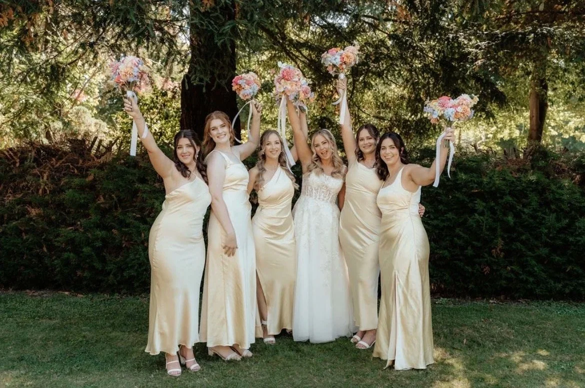 Bride with five bridesmaids outdoors, holding bouquets, standing under trees.