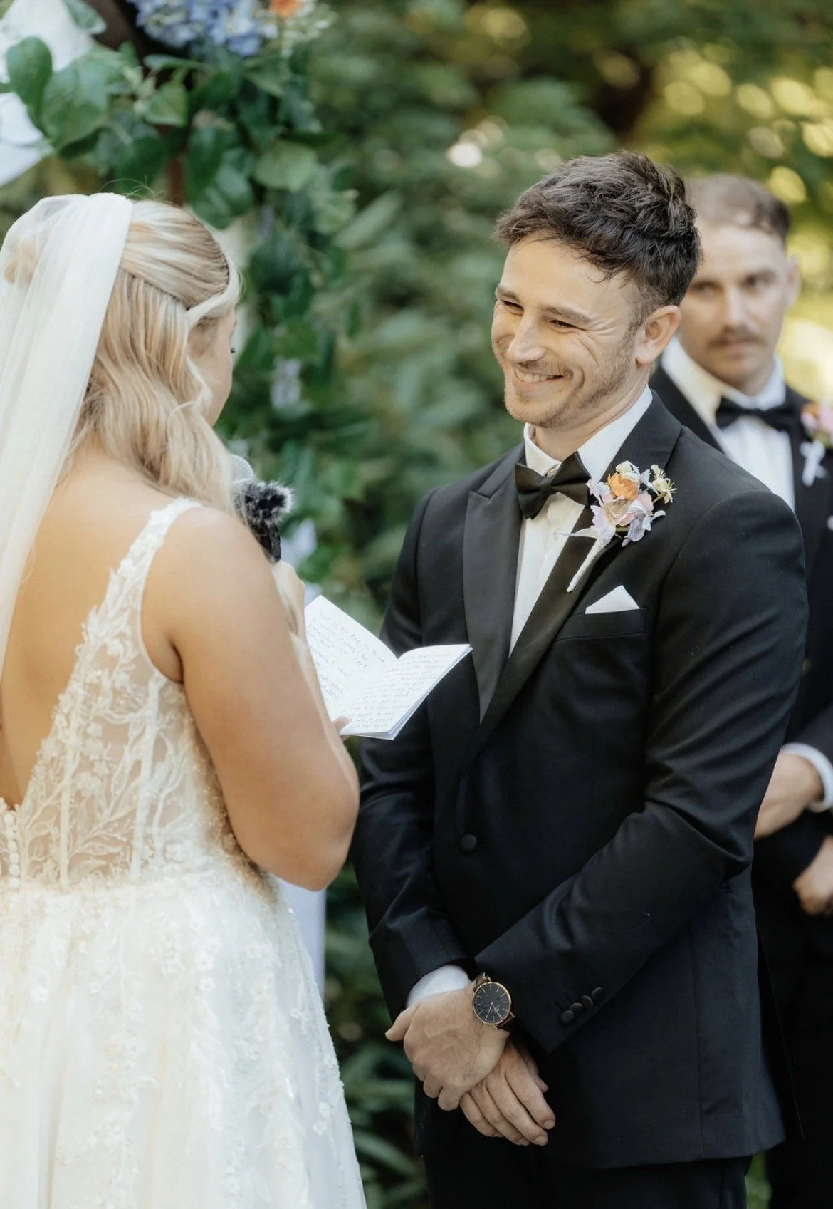 A wedding ceremony taking place outdoors with a bride and groom exchanging vows, with a groomsman in the background.