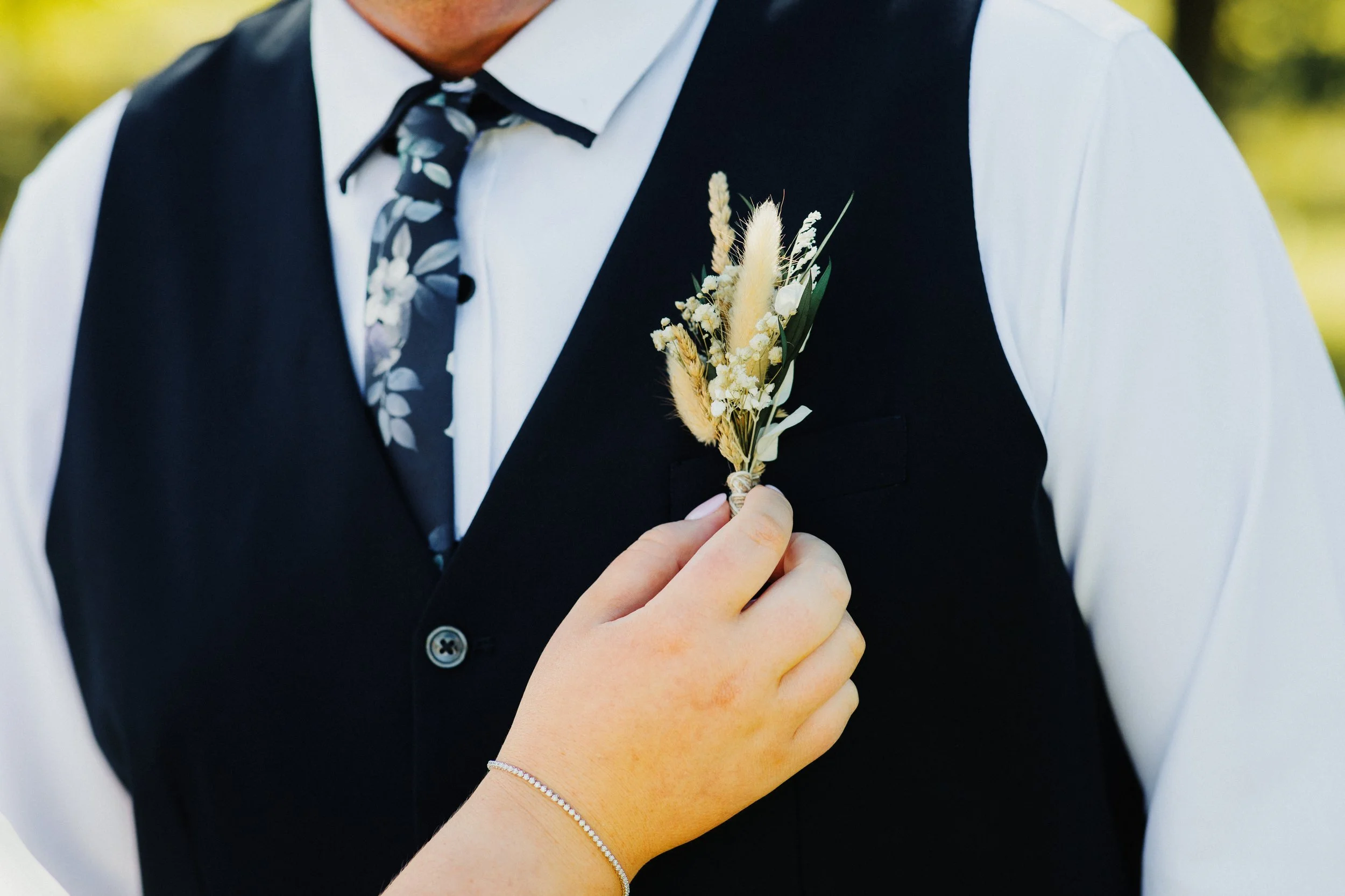 Person in formal attire with a white shirt, dark vest, and patterned tie, holding a small bouquet of dried flowers on their chest