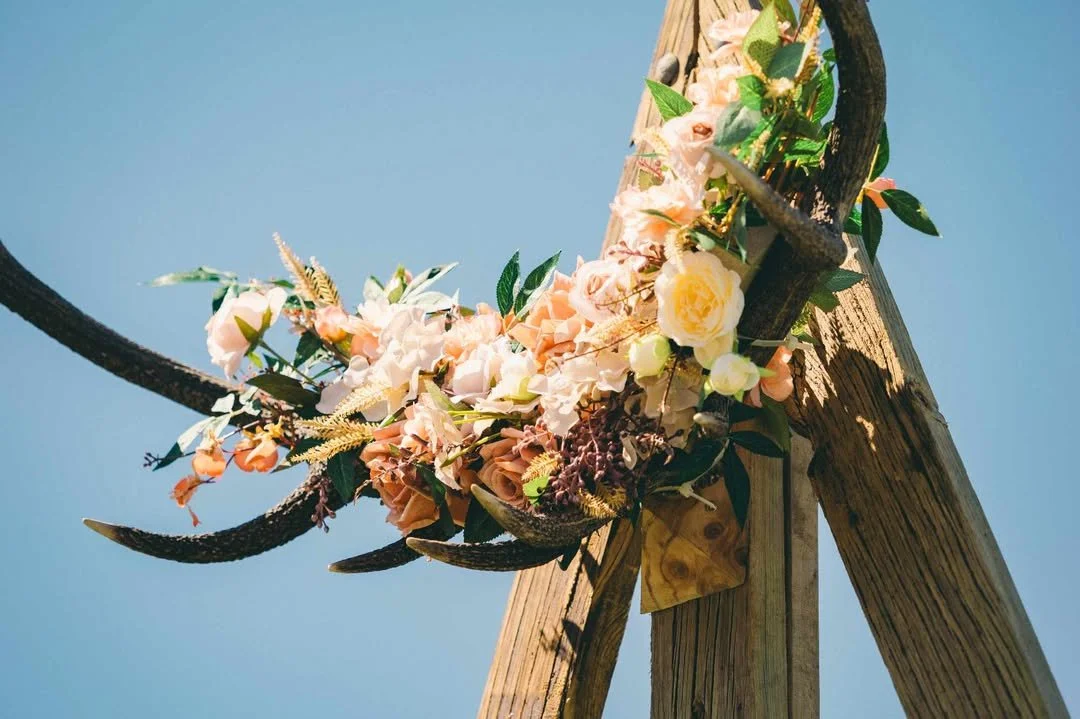 Decorative flower arrangement with pink and white flowers and dark animal horns on a wooden stand against a blue sky