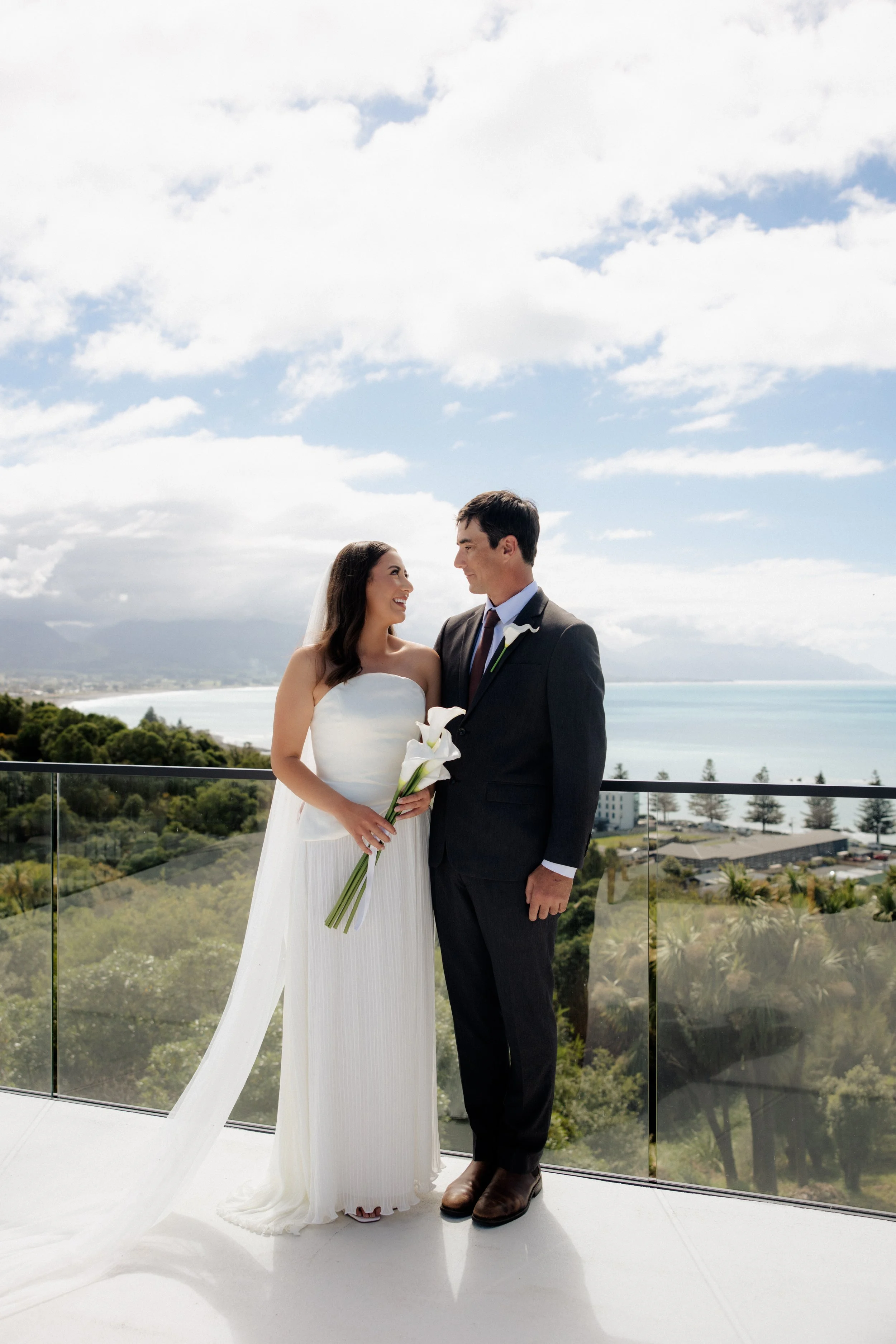 A bride and groom standing on a balcony with a scenic view of the ocean and sky in the background. The bride is wearing a strapless white wedding dress and holding a small bouquet of calla lilies, while the groom is dressed in a dark suit with a tie. They are smiling at each other.