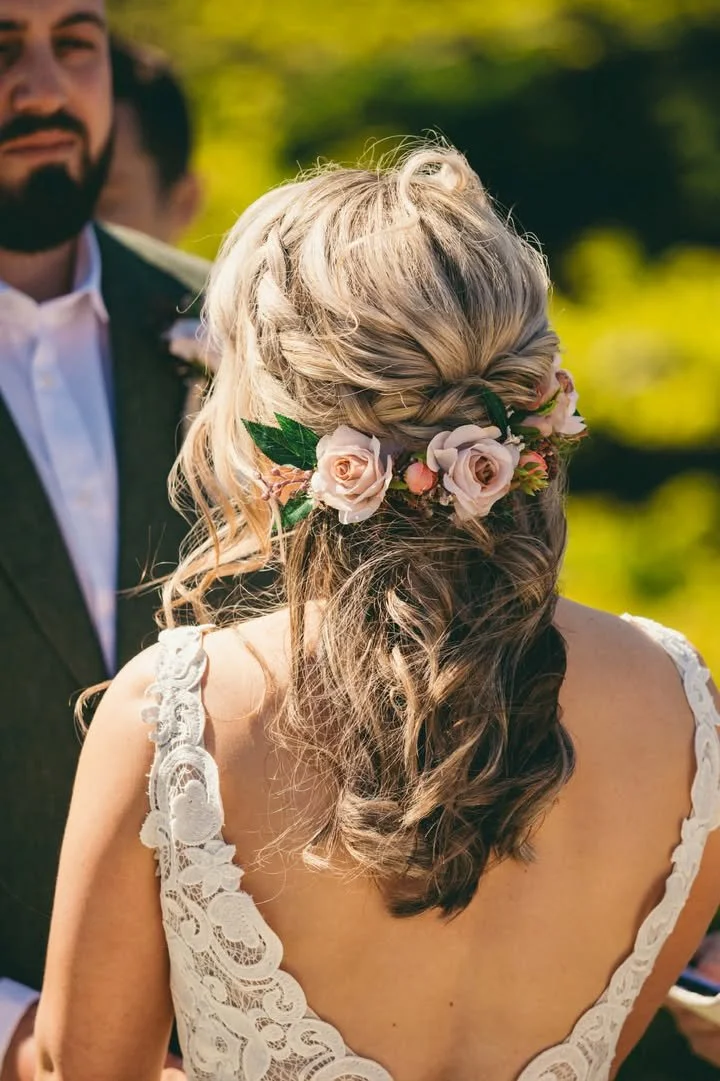 Back of a bride with wavy hair wearing a floral crown of pink roses and greenery at an outdoor wedding with a groom in a suit in the background.