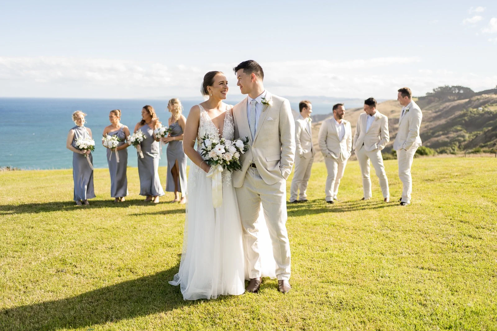 A bride and groom holding a bouquet looking at each other while standing on a grassy area near the ocean during a seaside wedding. Bridesmaids and groomsmen stand in the background.