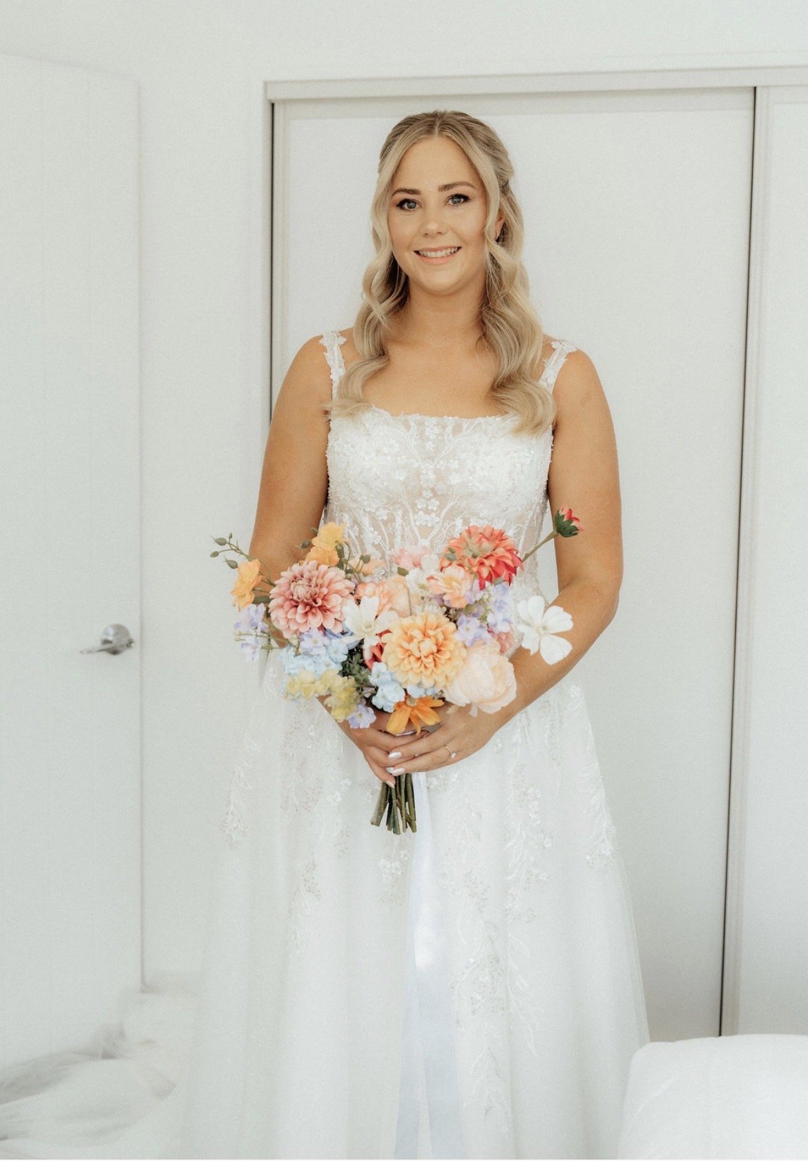 A smiling woman in a white wedding dress holding a bouquet of colorful flowers indoors.