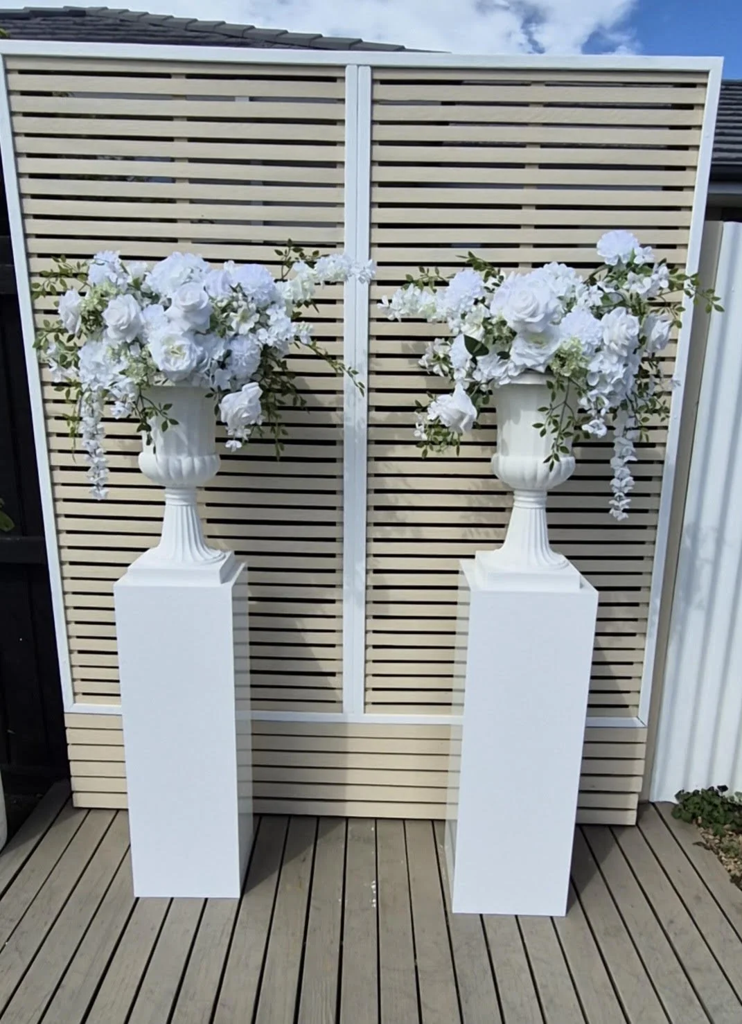 Two white pedestals with large white floral arrangements in ornate white vases, positioned in front of a decorative wooden privacy screen on a wooden deck.