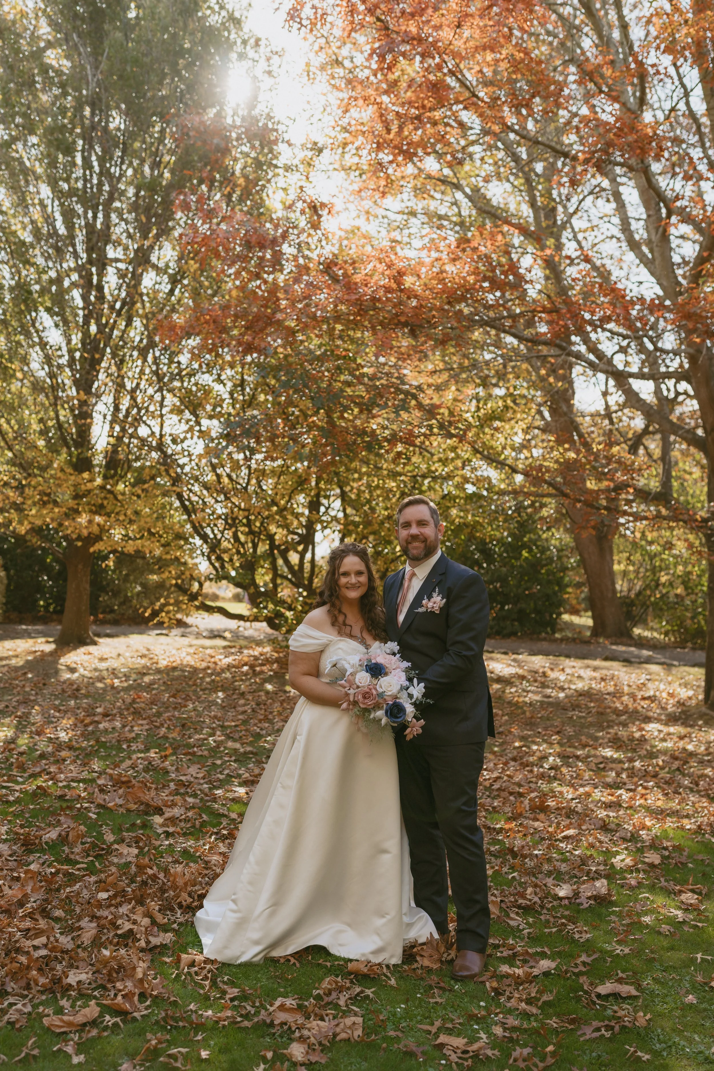 Autum trees and leaves on the ground at golden hour of Bride and Groom posing together - bride with her bouquet