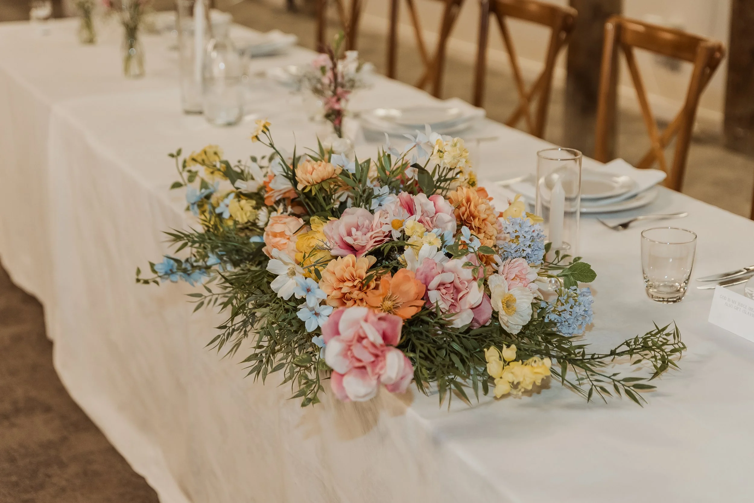 A wedding table centerpiece with a large, colorful bouquet of flowers including pink, peach, yellow, and blue blooms, set on a cream tablecloth with table settings and glasses, in a rustic indoor setting.