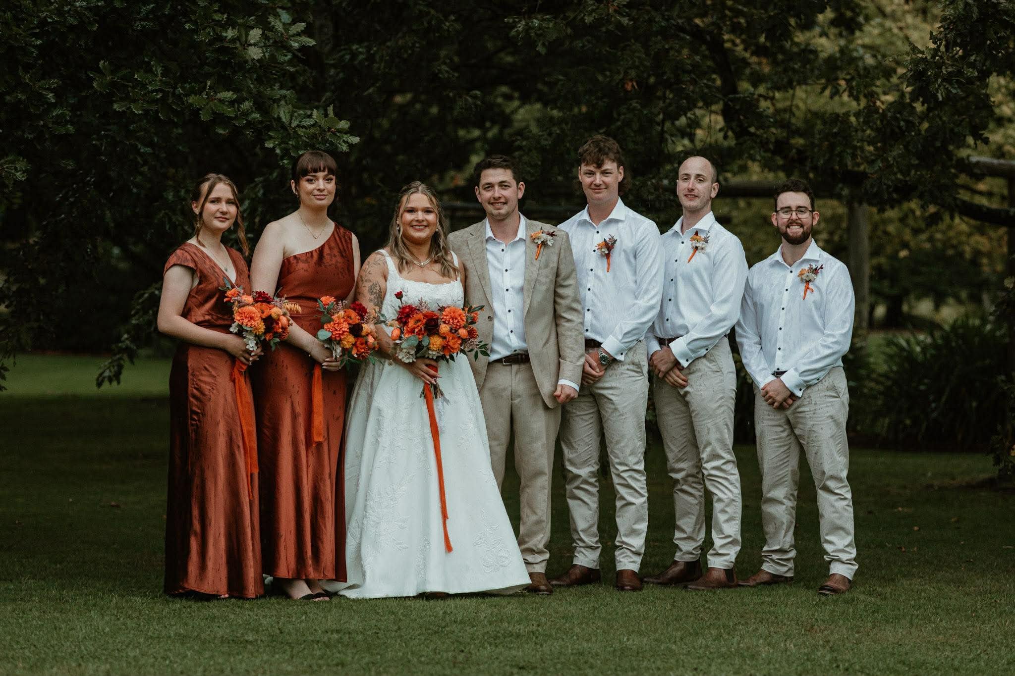 A wedding party with six people standing outdoors on grass, dressed in formal attire, with three women holding bouquets of orange and red flowers, and three men wearing white shirts with boutonnieres. They are posed in front of a background of green trees.
