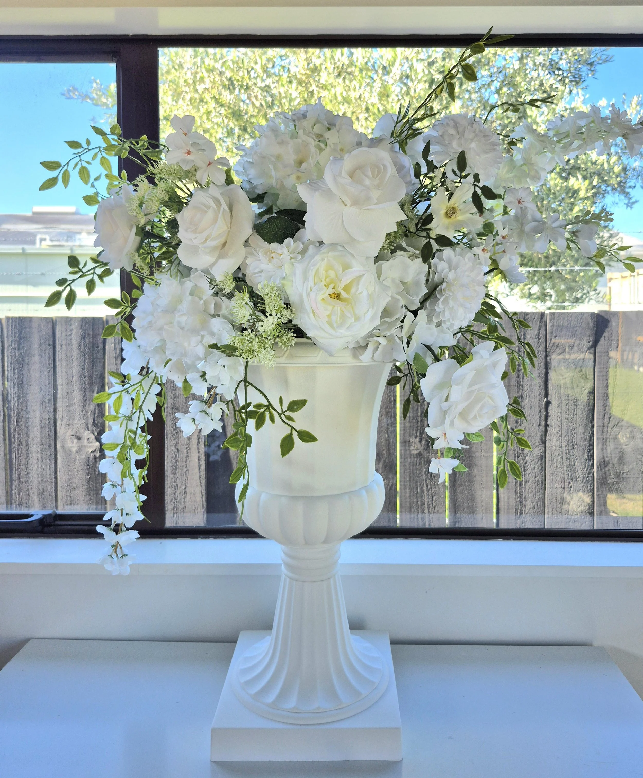A white floral arrangement in an elegant white vase, with a mix of white roses, hydrangeas, and greenery, placed on a white surface in front of a window.