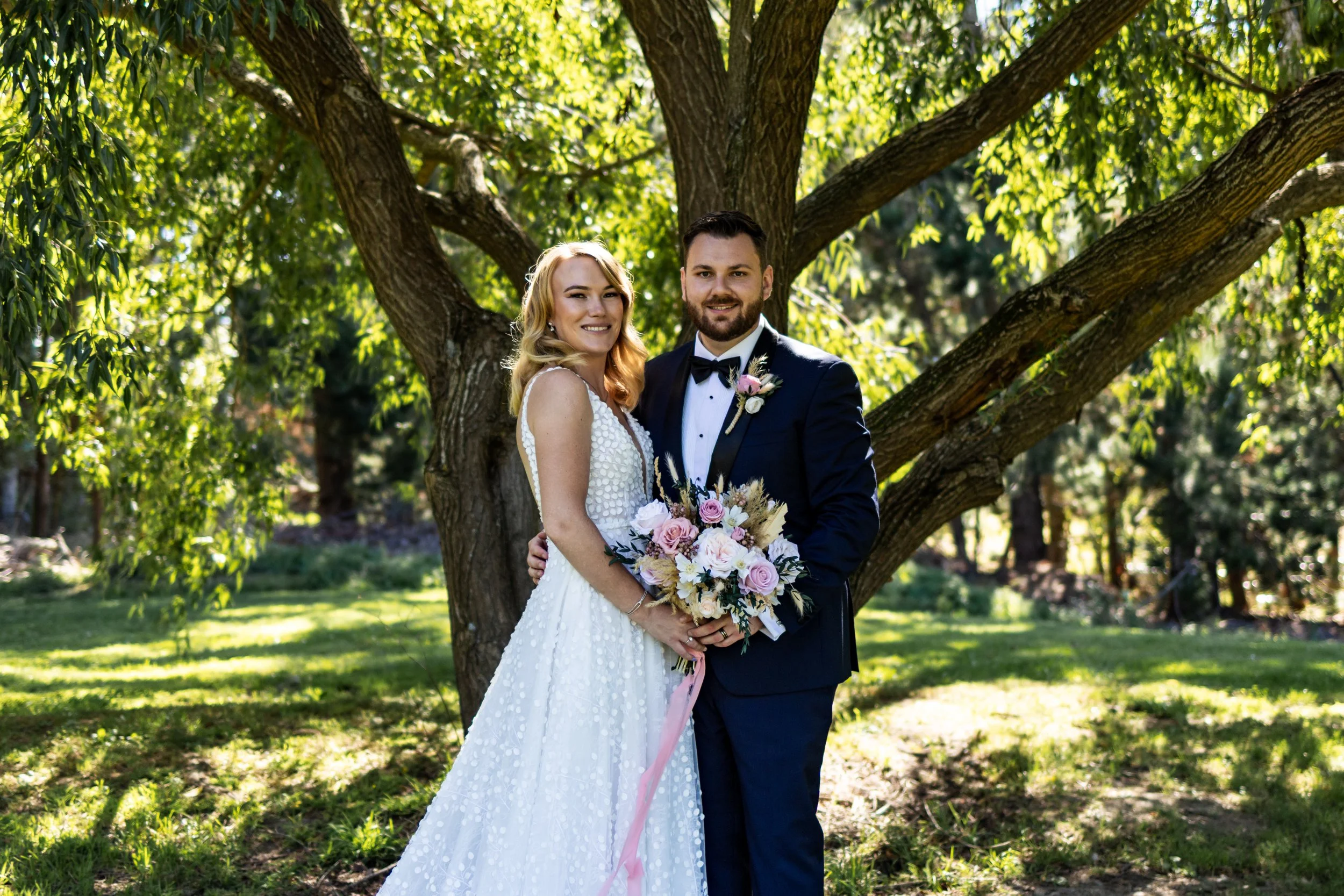 A bride and groom standing outdoors under a large tree on a sunny day, smiling for a wedding photo. The bride is holding a bouquet, wearing a white dress, and the groom is in a black tuxedo with a bow tie.
