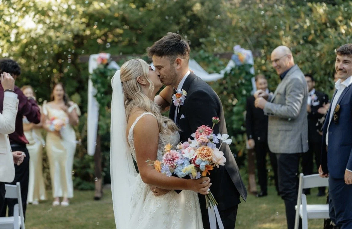 Bride holding her colourful bouquet at the end of the aisle whilst groom holds and kisses her
