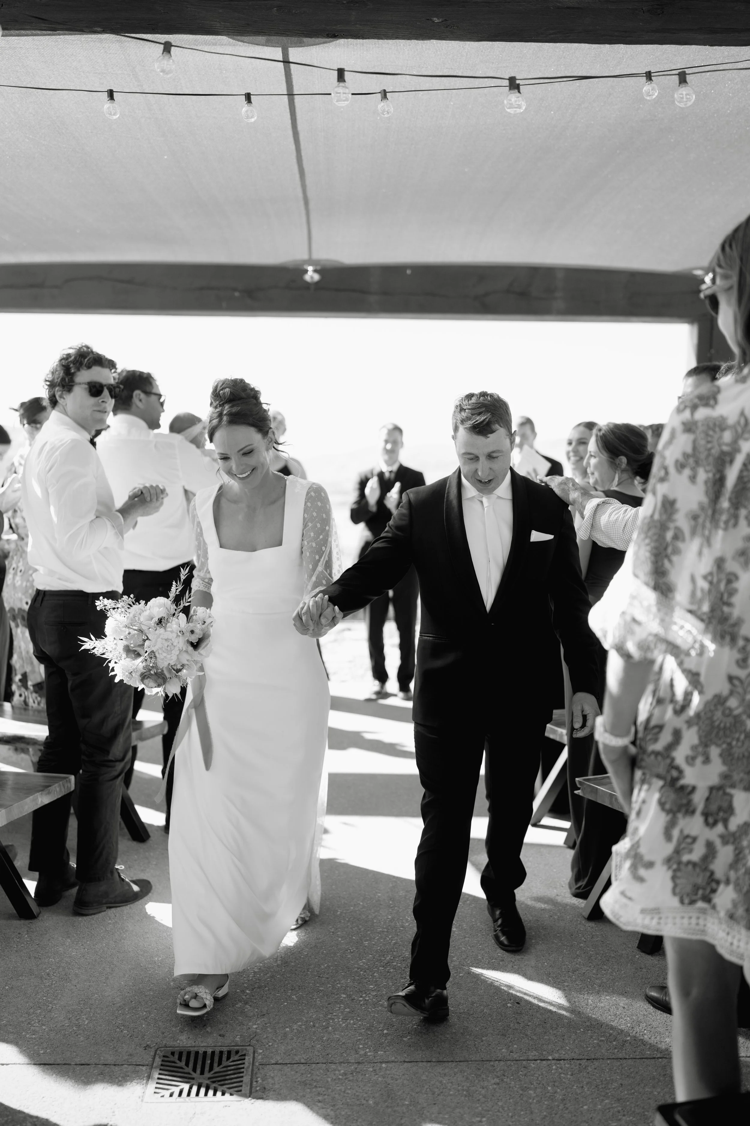 Black and white photo of a wedding ceremony. The bride and groom are walking hand in hand in the center, smiling. The bride is wearing a white dress and holding a bouquet, the groom is in a dark suit. Guests are standing on either side, some clapping and smiling. Overhead string lights are hanging under a shaded canopy.