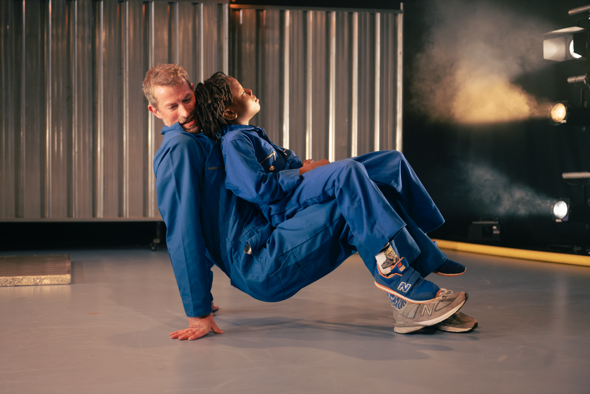 A white man and a Black boy are playing together. The boy is sitting on the man and he walks on hands and feet across the stage. They are both dressed in blue jumpsuits. The floor is blue and they are in front of a silver screen.