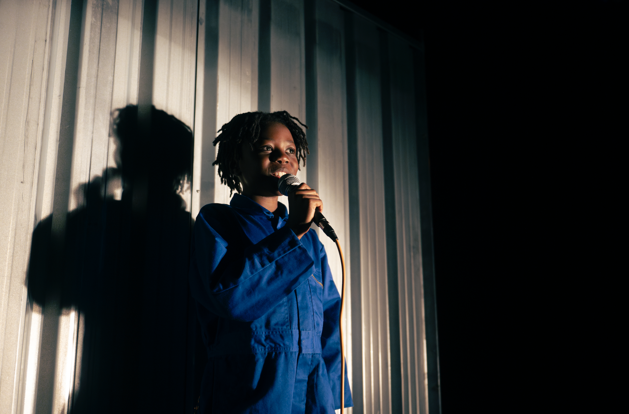 A Black young boy with dreadlocks wearing a blue jumpsuit holding a microphone that he is singing into. His shadow is cast on the metal wall behind him.