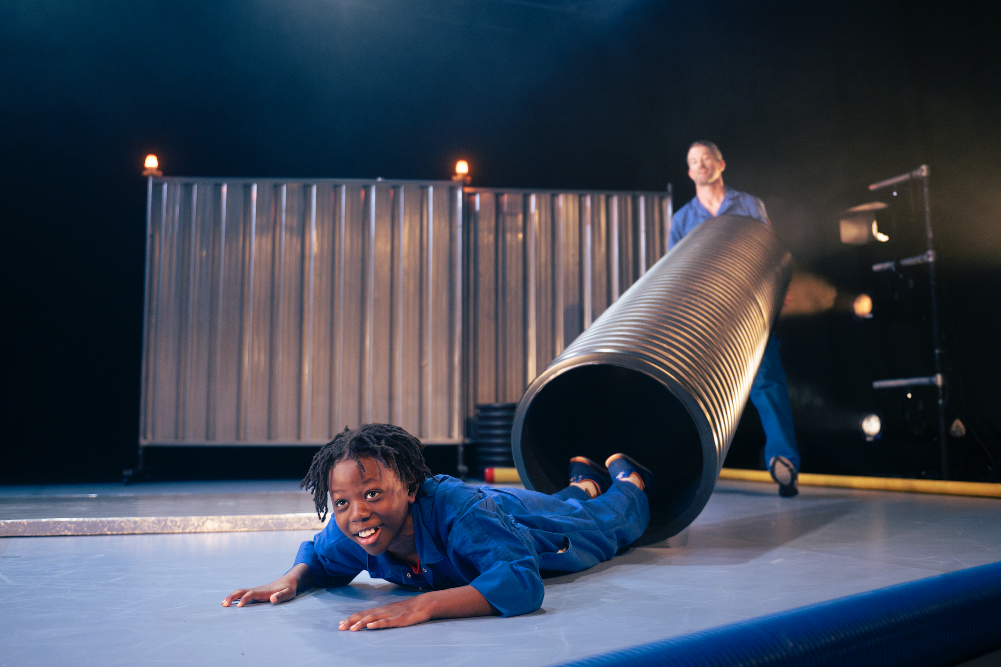 A Black boy in a blue jumpsuit sliding out of a large black pipe on a stage, with a man standing behind the pipe, lifting it up and smiling. The stage has a blue floor, metallic screens on wheels and is lit with stage lights.