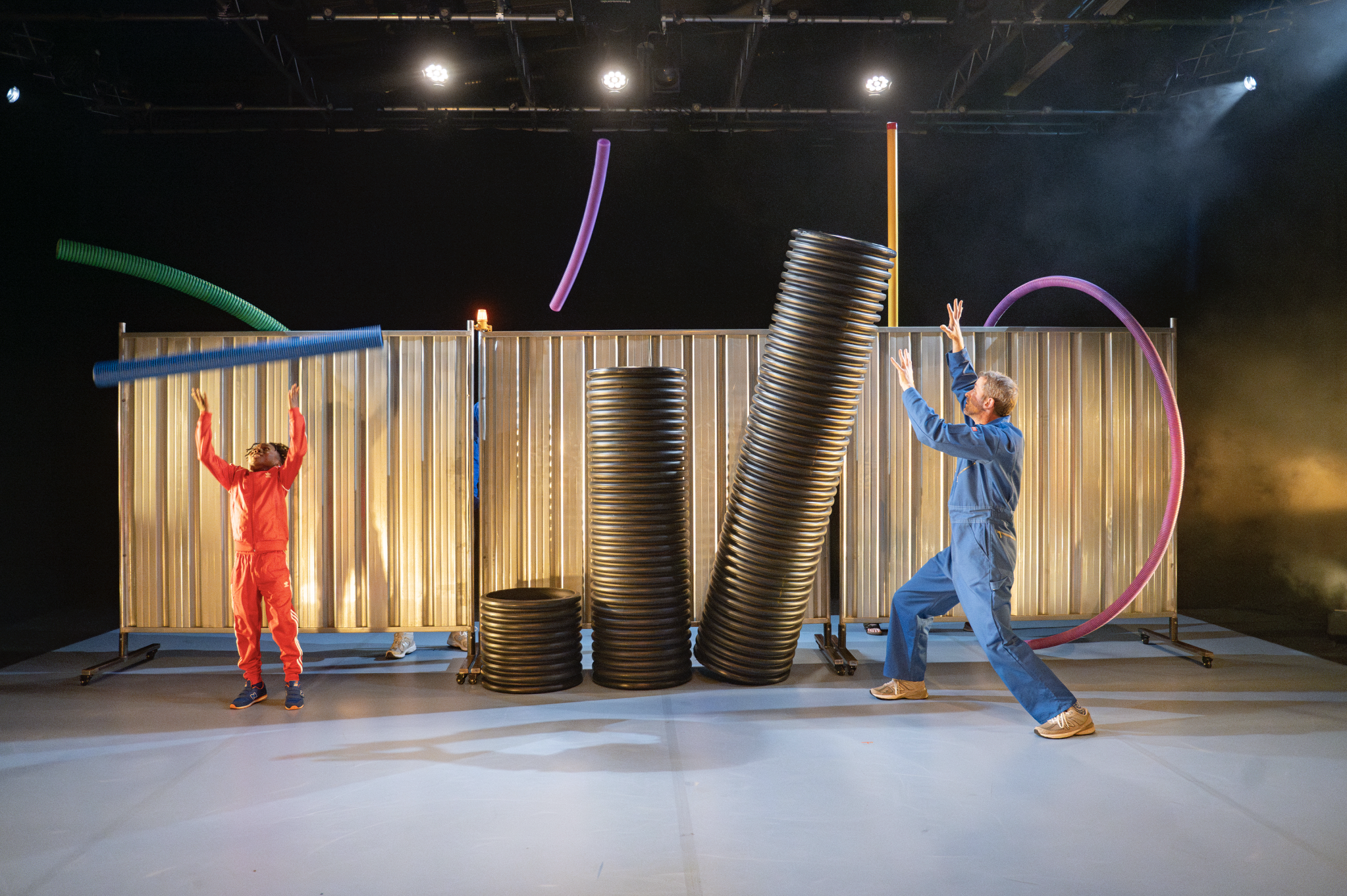 Two performers -  a Back boy in a red adidas tracksuit and an older man in a bright blue jumpsuit catching colourful plastic pipes as they're thrown over  a metal barrier.