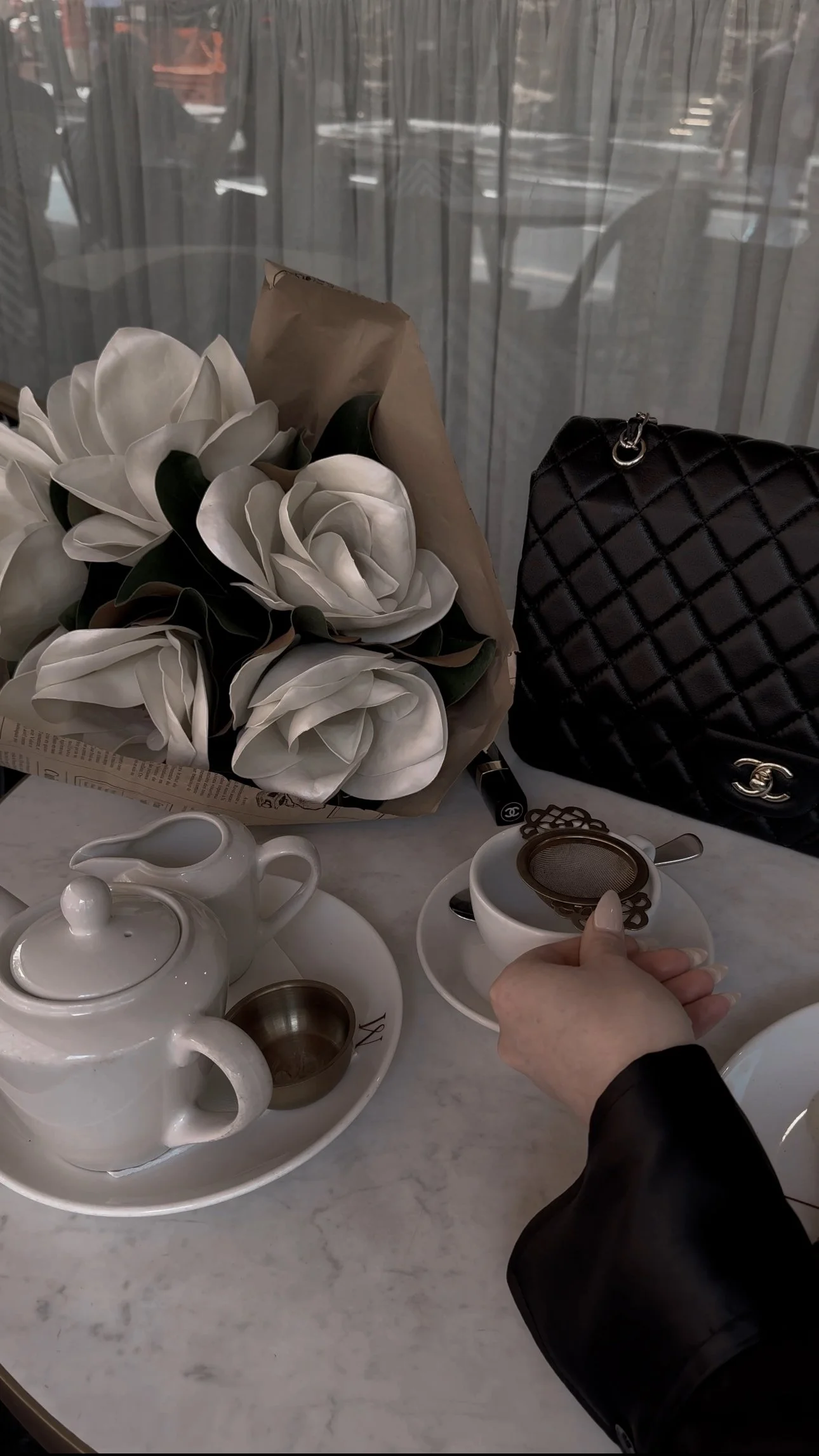 A marble table with a bouquet of white roses, a teapot with a sugar bowl, a cup of tea with a tea infuser, a Chanel handbag, and some small items including lipstick and keys.