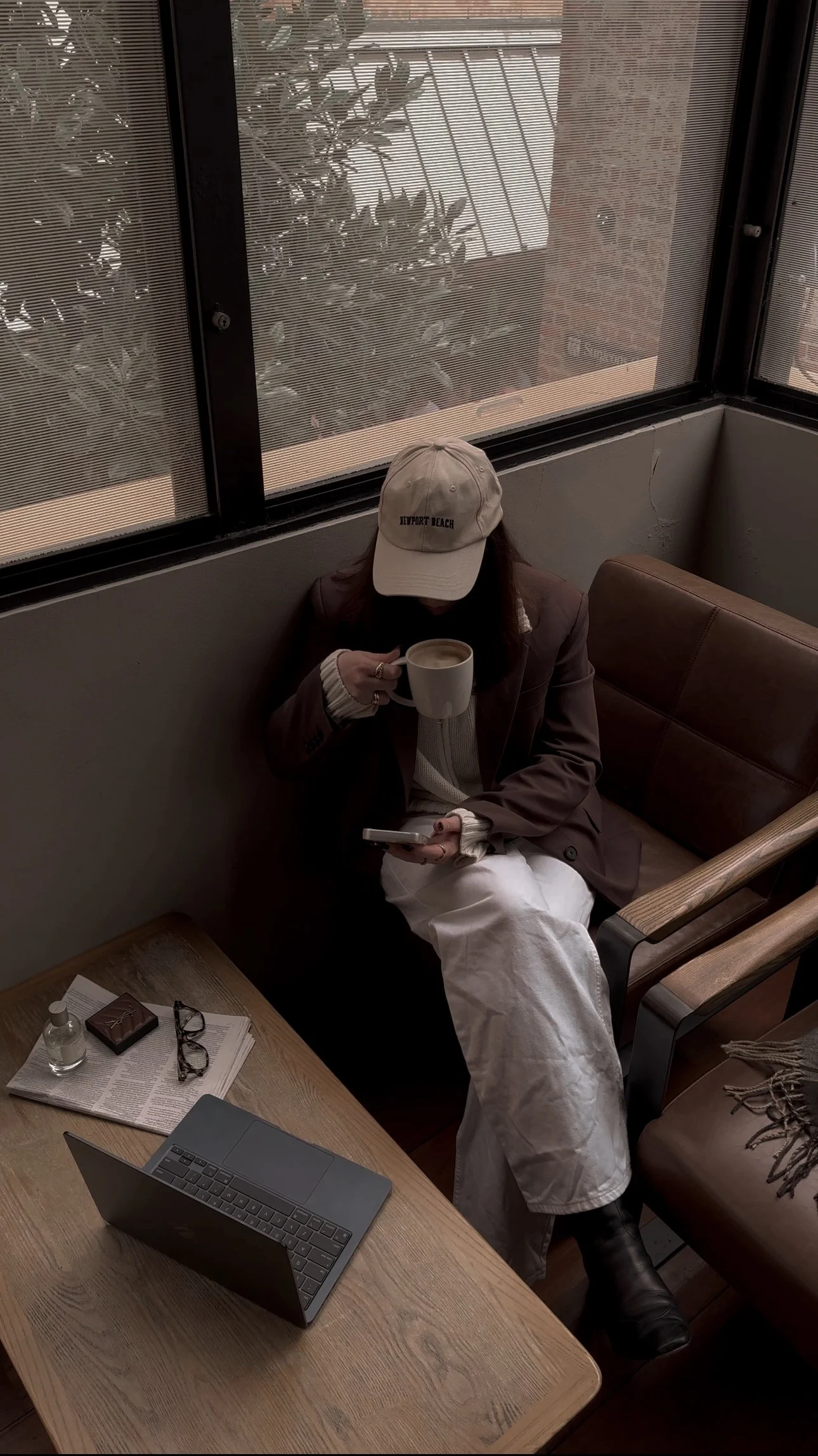 A woman sitting on a leather bench holding a coffee mug and a smartphone in a cozy cafe. She is wearing a beige cap, a brown blazer, and white pants. There are glasses, a notebook, a small bottle, and a laptop on the wooden table in front of her.