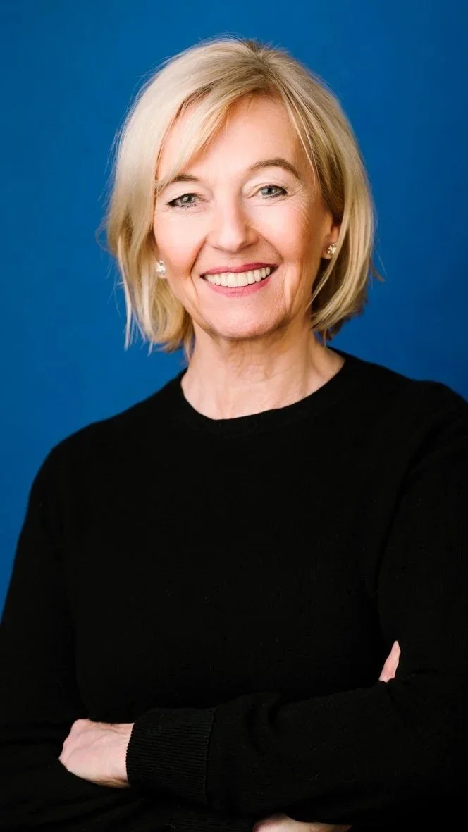 A smiling woman with blonde hair wearing a black top, standing against a blue background.