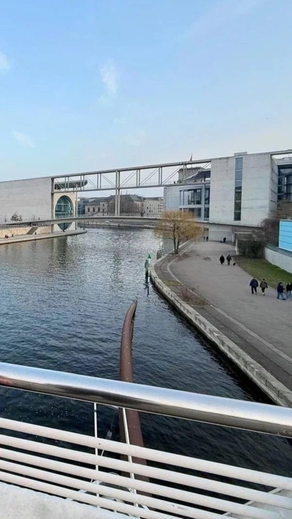 Modern cityscape with a canal, pedestrian walkway, and contemporary architecture, including a bridge and large glass building.