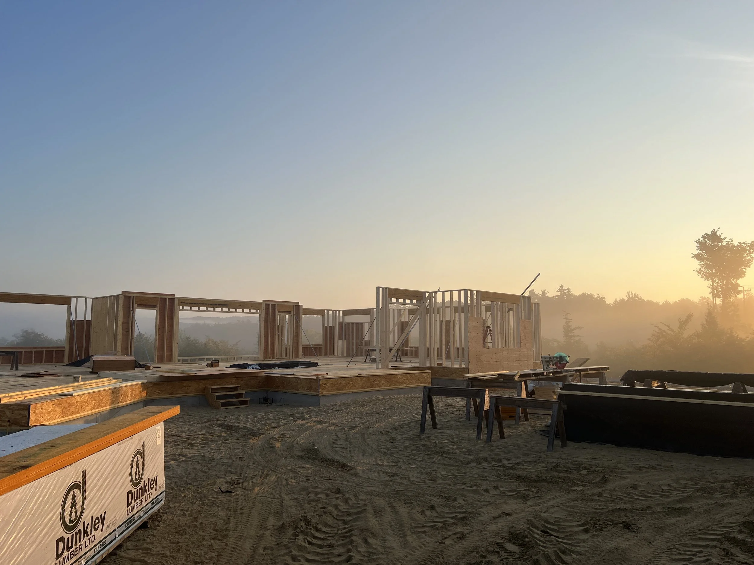 Construction site during sunrise with wooden framing for a building, construction materials, and tools scattered around the dirt ground.