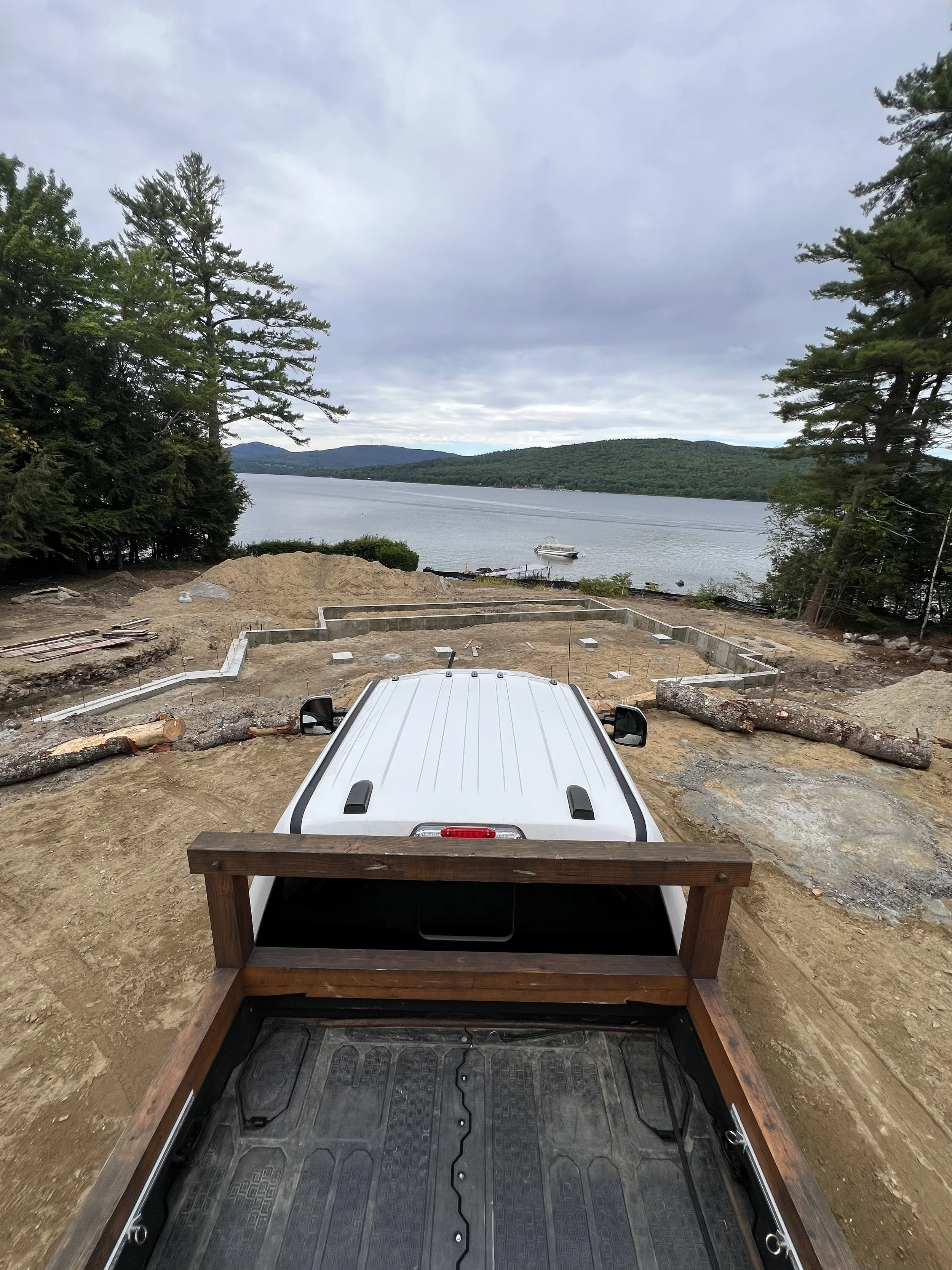 View from a pickup truck bed pointing towards a lakeside construction site with trees, sand, and a boat in the water under cloudy sky.