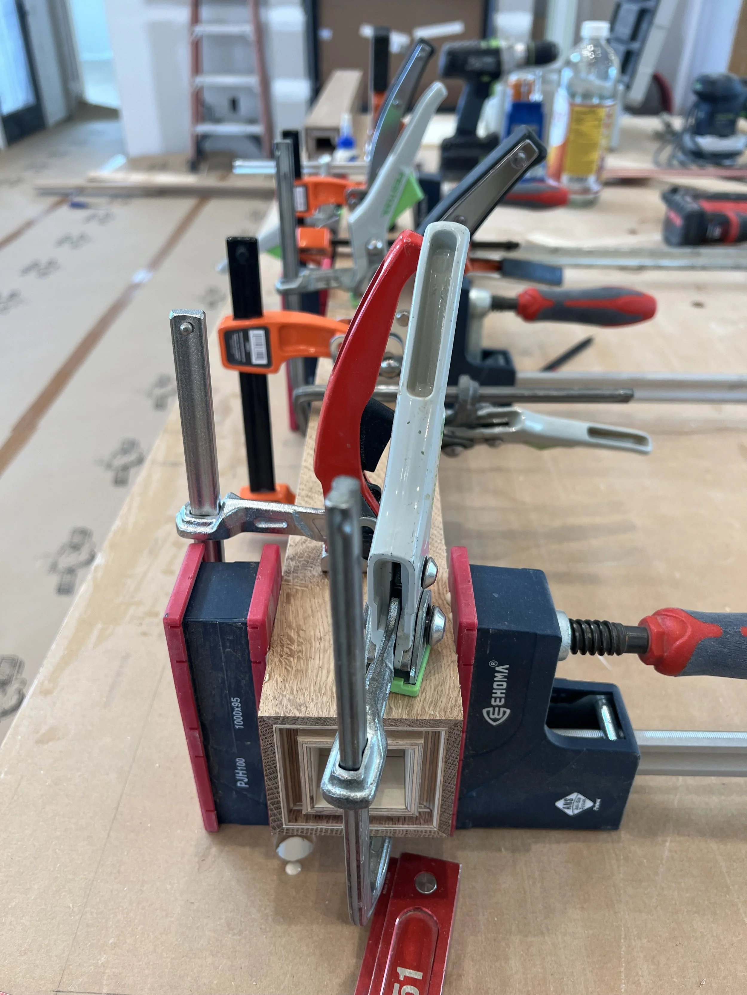 Clamps holding wood pieces with a detailed recessed design in place on a workbench, surrounded by woodworking tools and equipment.