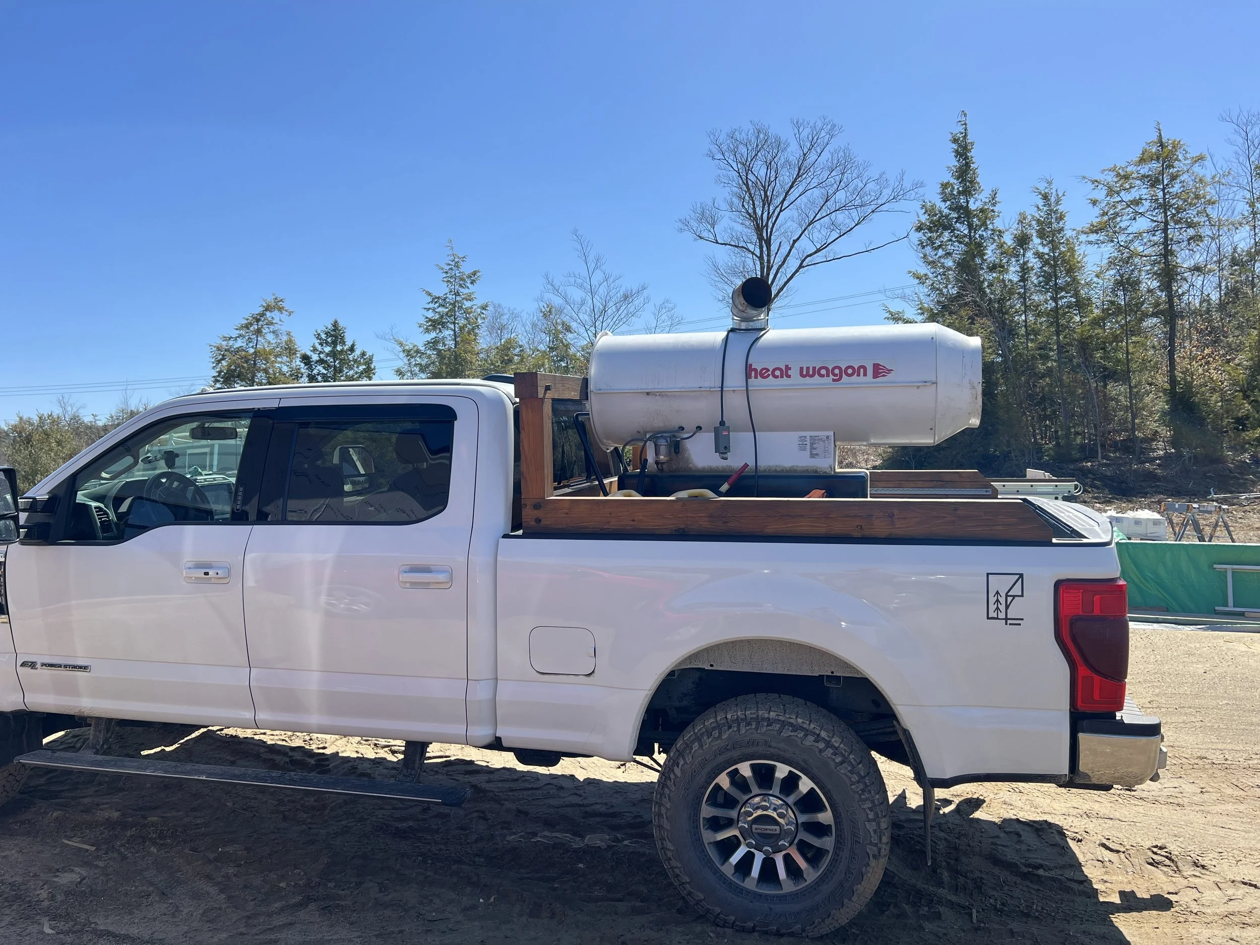 A white pickup truck with a wooden bed and a large cylindrical tank labeled 'heat wagon' mounted on the back, parked on a dirt lot beneath a clear blue sky and surrounded by trees.