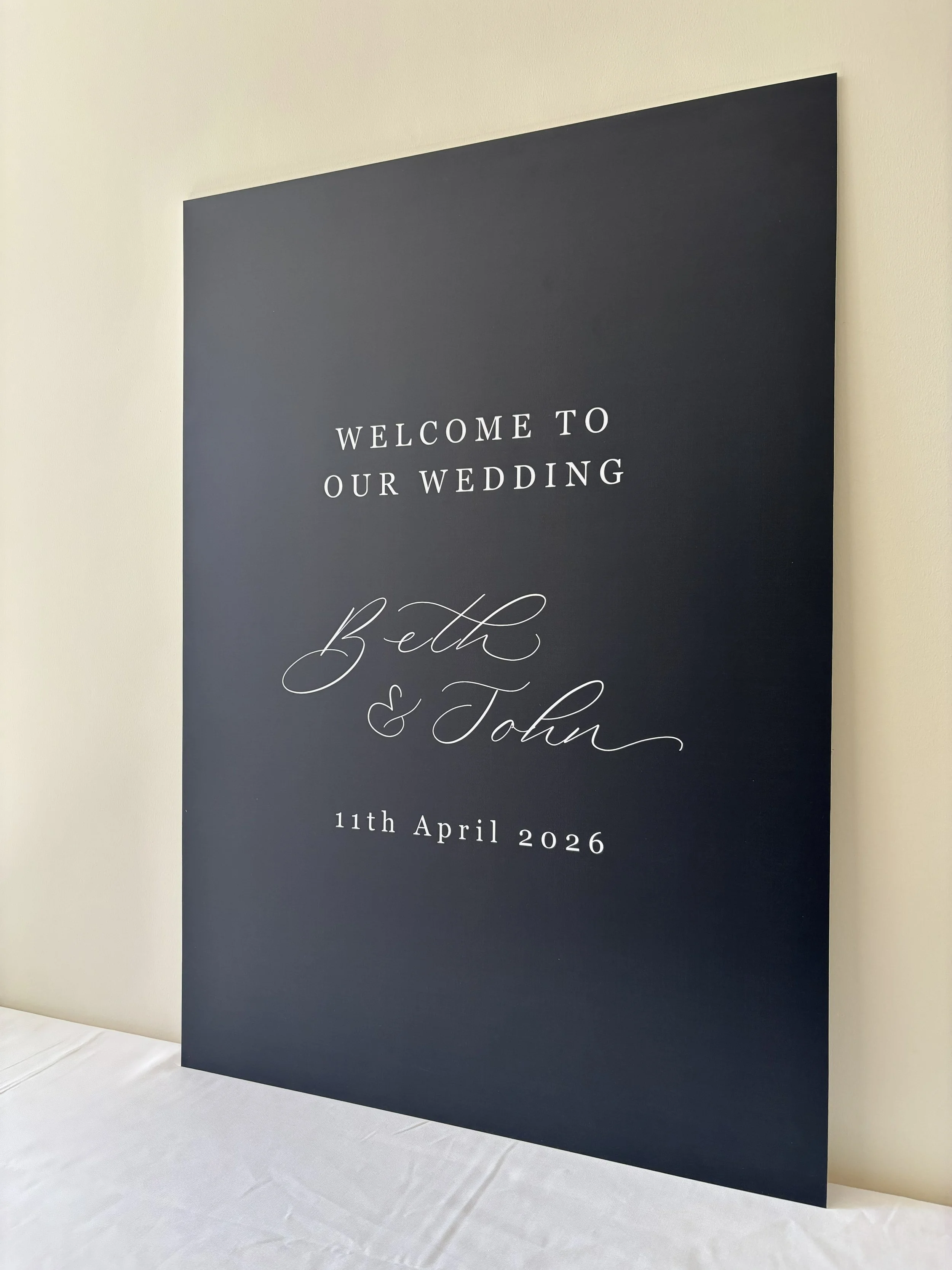 An A1, dark navy wedding welcome sign leaning against a light-colored wall. The sign features elegant white modern calligraphy that reads “Welcome to our wedding,” followed by the names “Beth & John”, and the date “11th April 2026” below. 