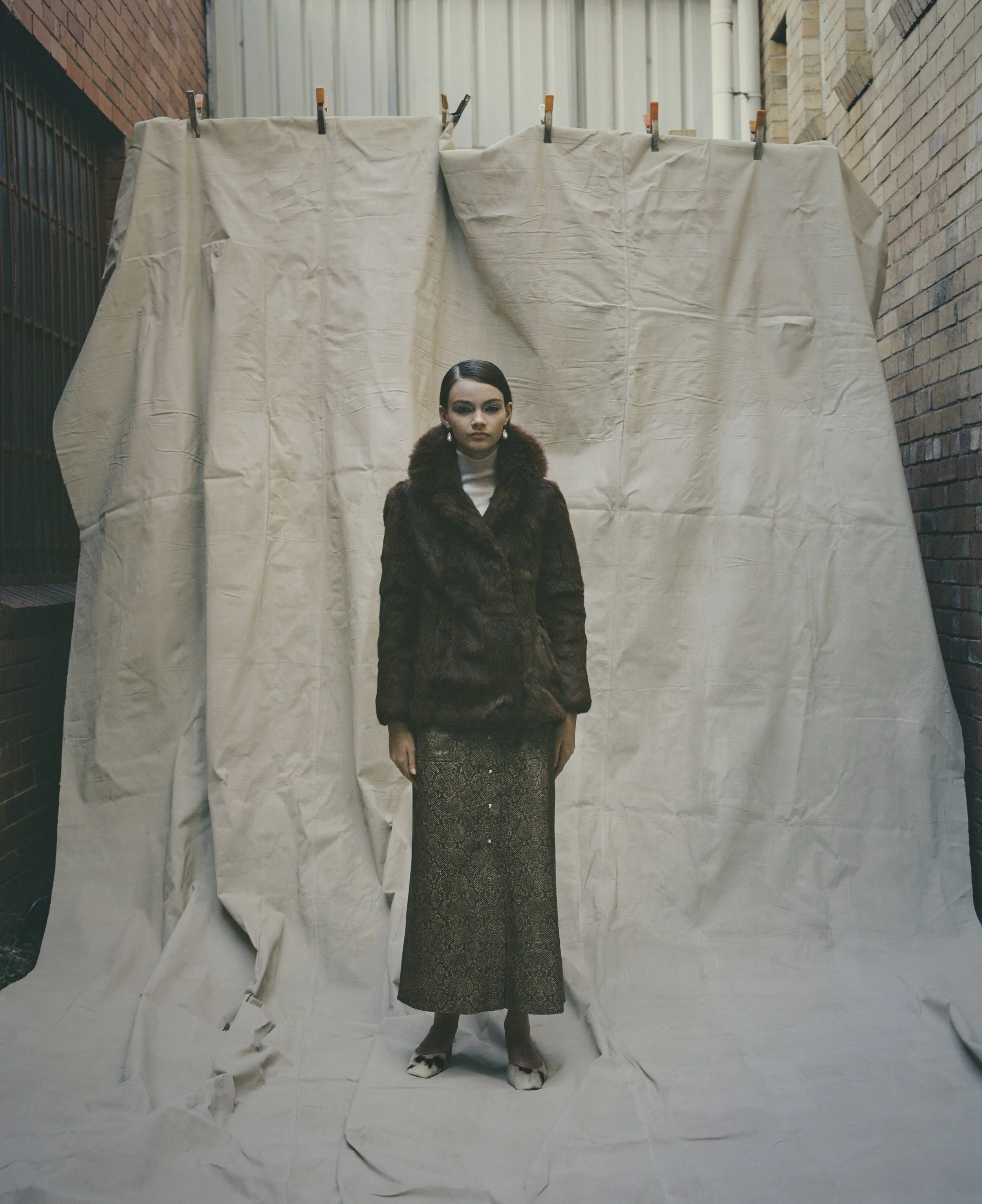 A woman stands in front of a beige fabric backdrop, wearing a vintage brown fur coat, a cream turtleneck sweater by Courtney Zheng, a long snake-skin patterned skirt by Nanushka, and mango shoes, with industrial brick buildings on either side.