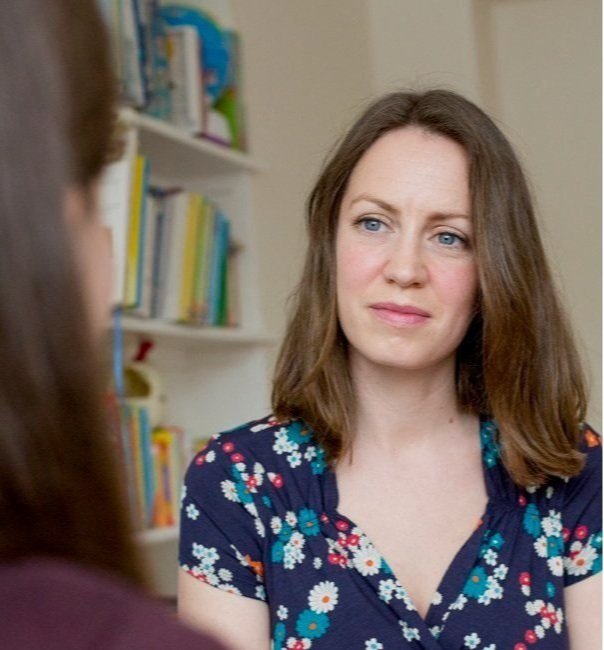 A woman with shoulder-length brown hair and blue eyes wearing a navy blue floral dress, sitting indoors with bookshelves in the background, engaged in conversation.