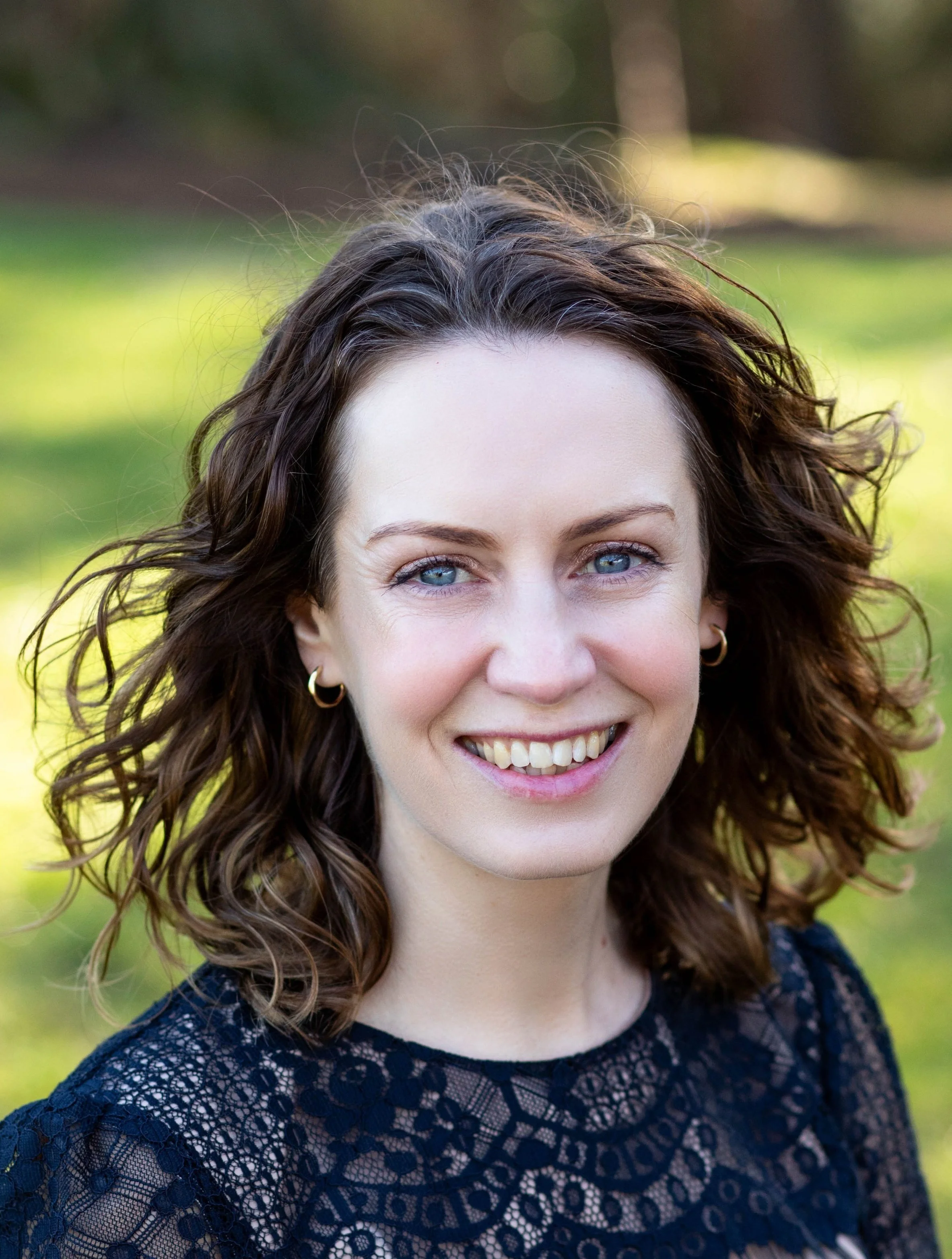 Close-up of a smiling woman with blue eyes and wavy brown hair outdoors in a park with green grass and trees in the background, wearing a navy blue lace top with hoop earrings.