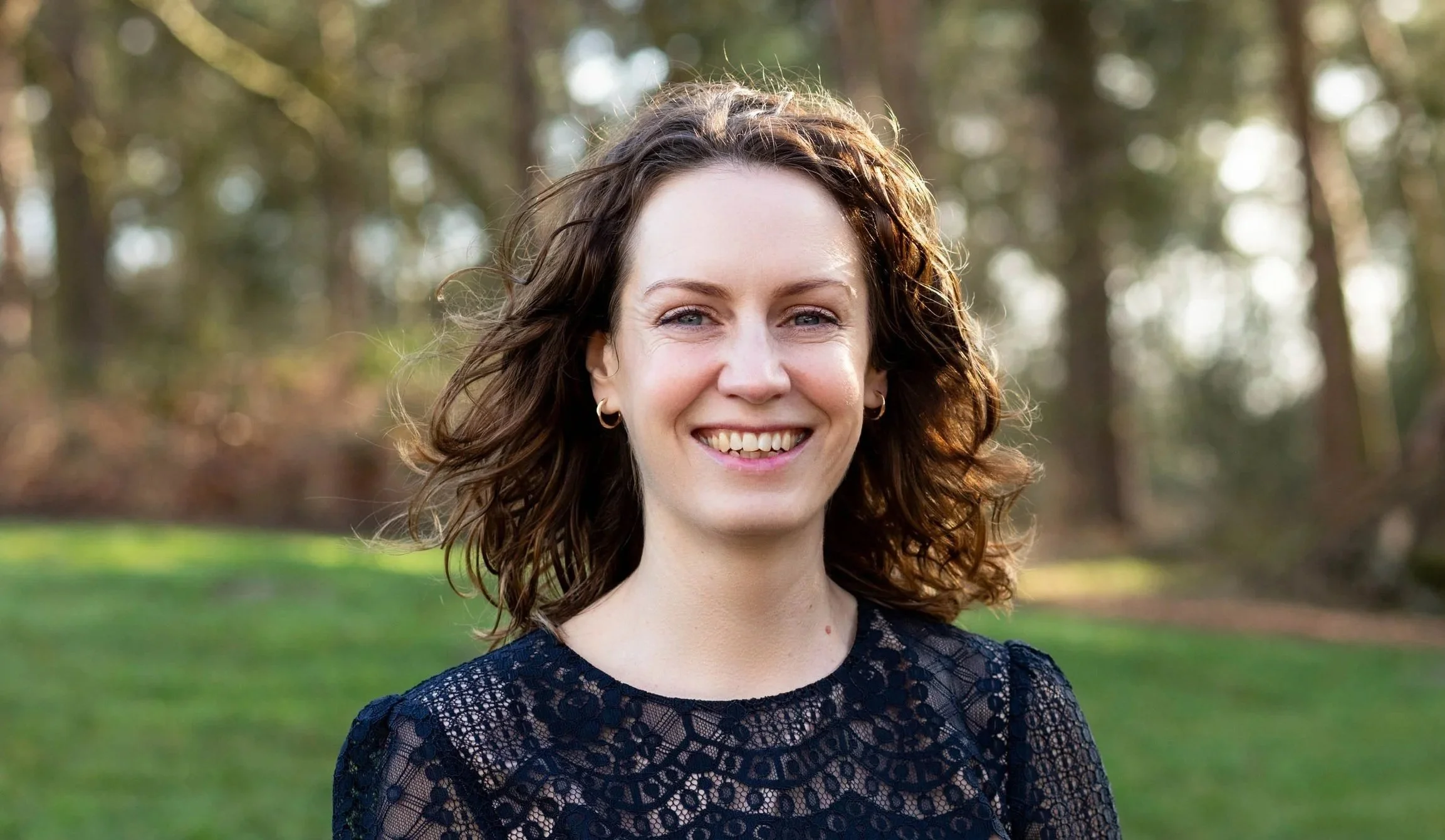 A woman with curly brown hair smiling outdoors in a park with green grass and blurred trees in the background.