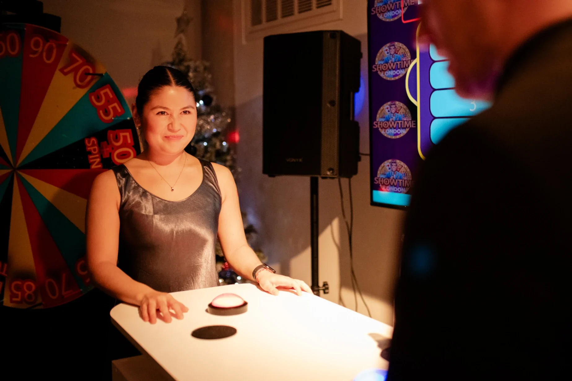 Woman standing behind a table with a button, smiling and looking at a person to her right. Behind her is a Christmas tree, a colorful wheel, and a screen with Showtime London logos.