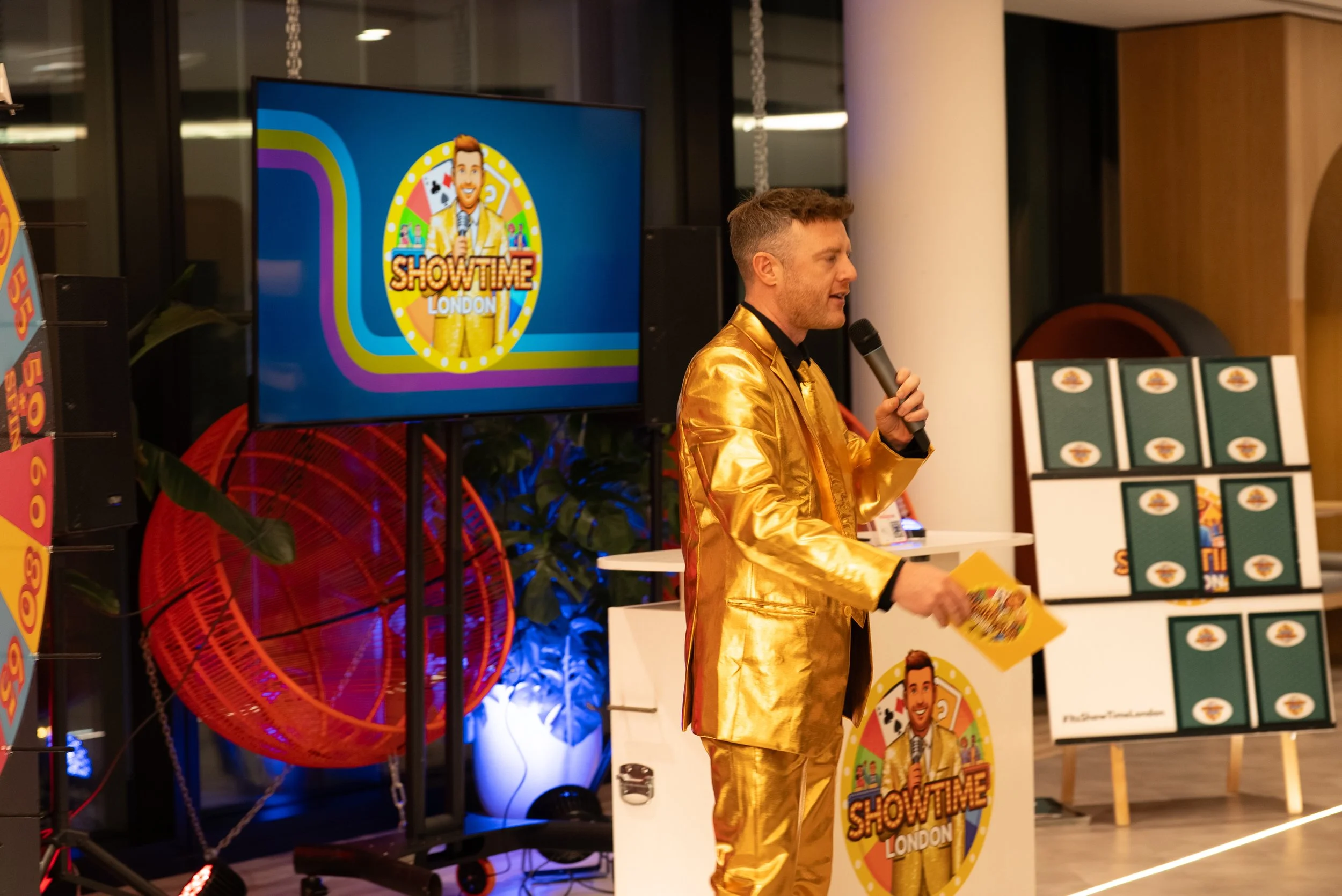 A man in a shiny gold suit holding a microphone, standing in front of a presentation screen with the Show Time logo, in an indoor event space.