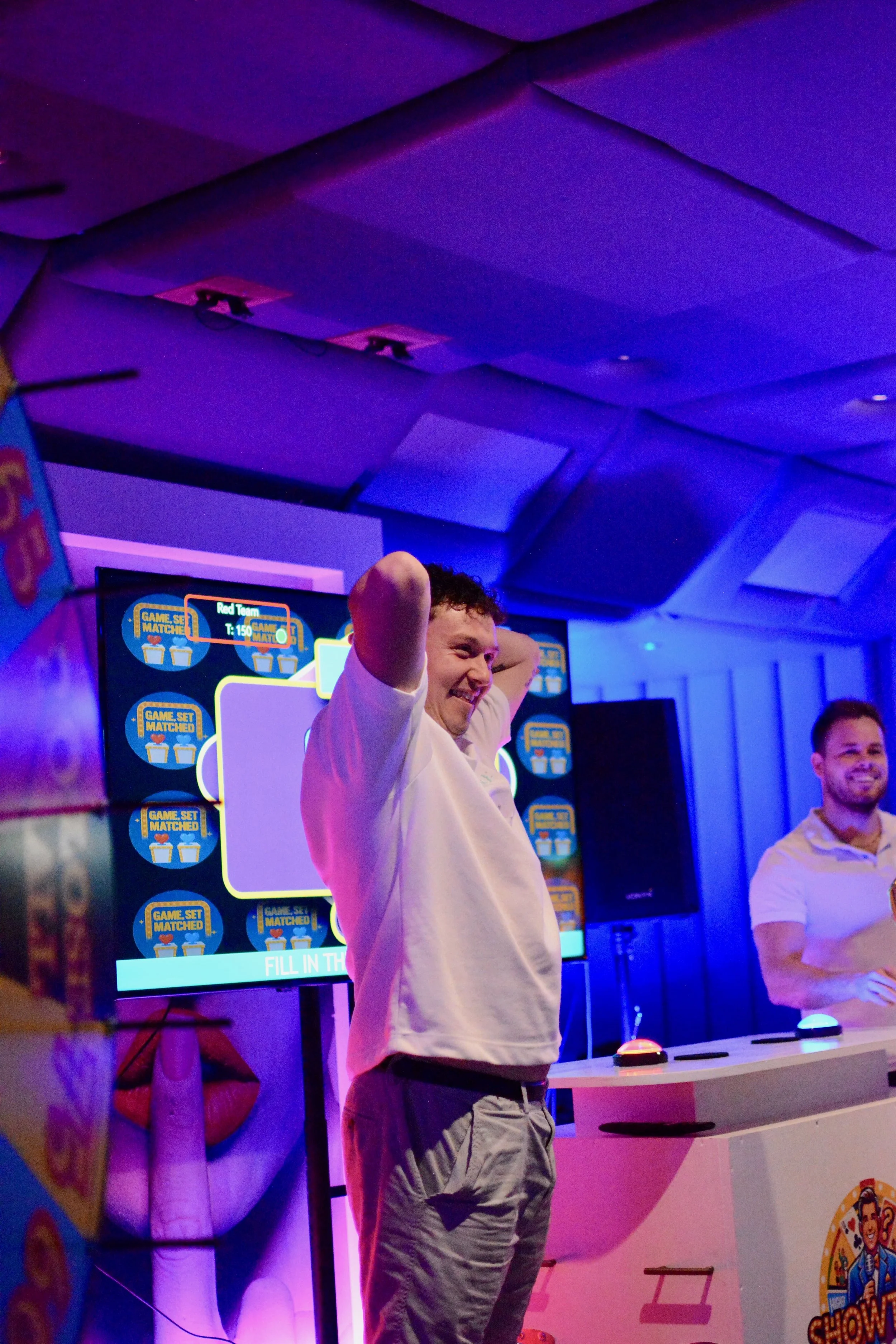 Two men are standing on a stage with a colorful game show background, featuring a large screen and illuminated buzzers. The man on the left is smiling, with his arms behind his head, while the man on the right is smiling with one hand on his hip, bot